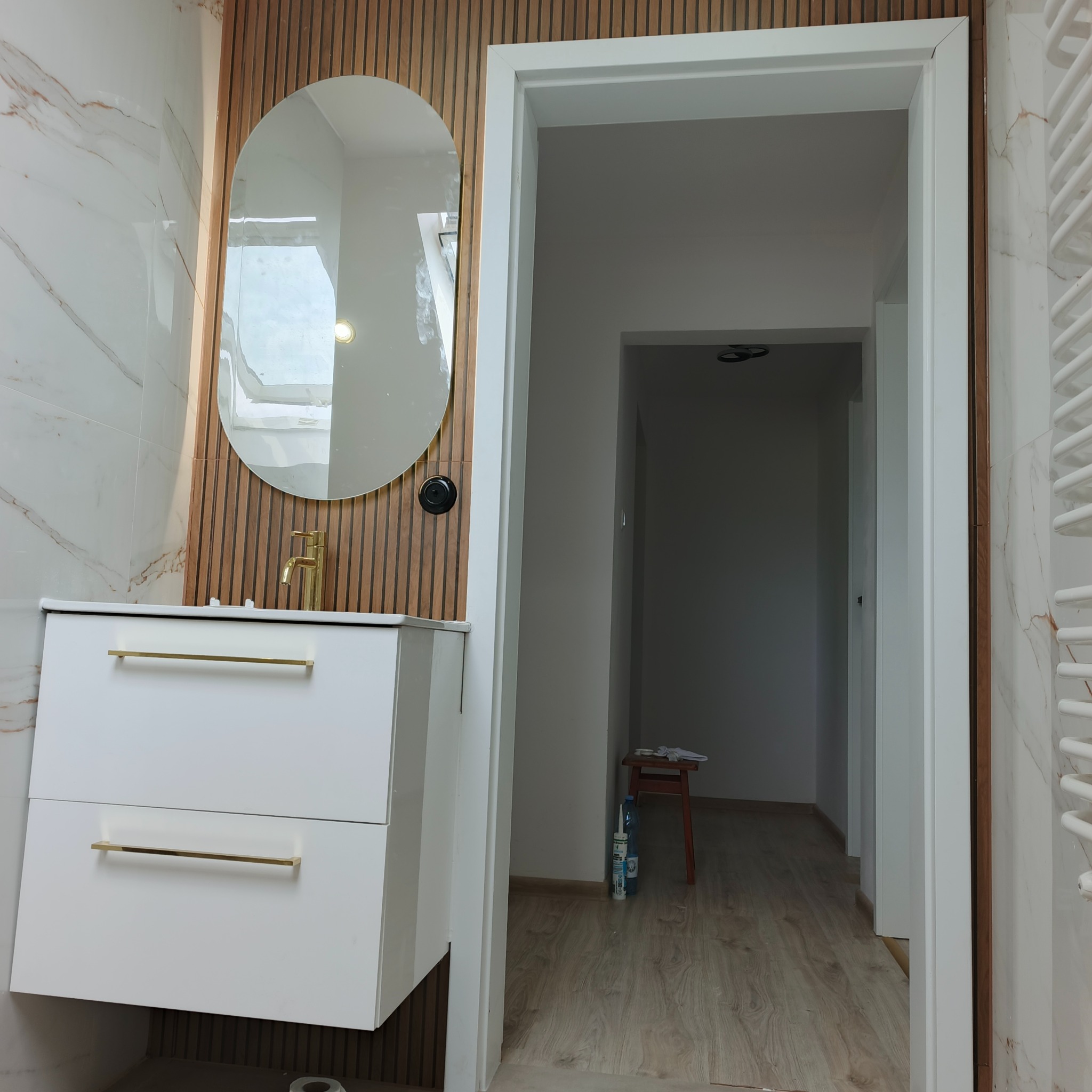 Nowoczesna łazienka z owalnym lustrem, białą szafką z złotymi uchwytami i drewnianymi lamelami na ścianie. Widok na korytarz z drewnianą podłogą.