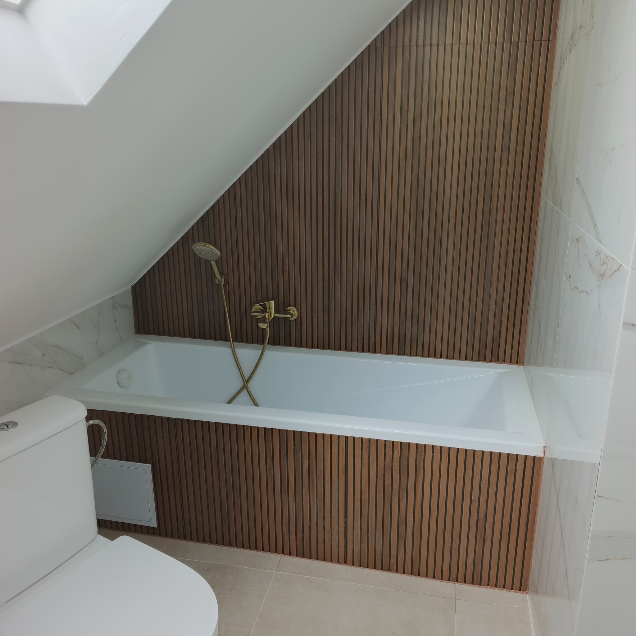 Biała wanna z złotą armaturą w łazience na poddaszu, wykończonej drewnianymi panelami ściennymi i marmurowymi płytkami. Widoczny fragment toalety.