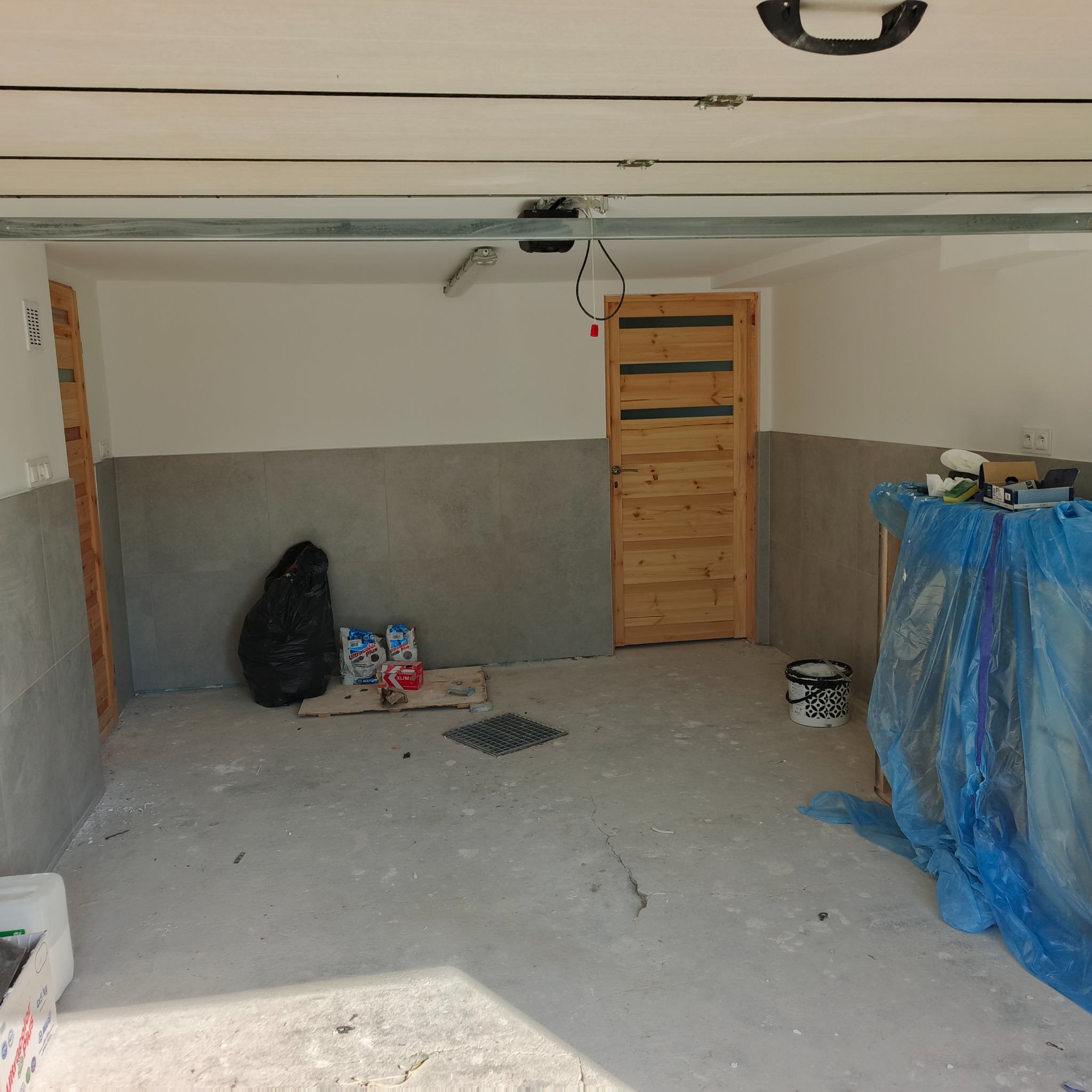 Wnętrze garażu po częściowym remoncie: ściany wyłożone szarą płytką do połowy wysokości, drewniane drzwi z przeszkleniami, worki z zaprawą, podłoga betonowa.