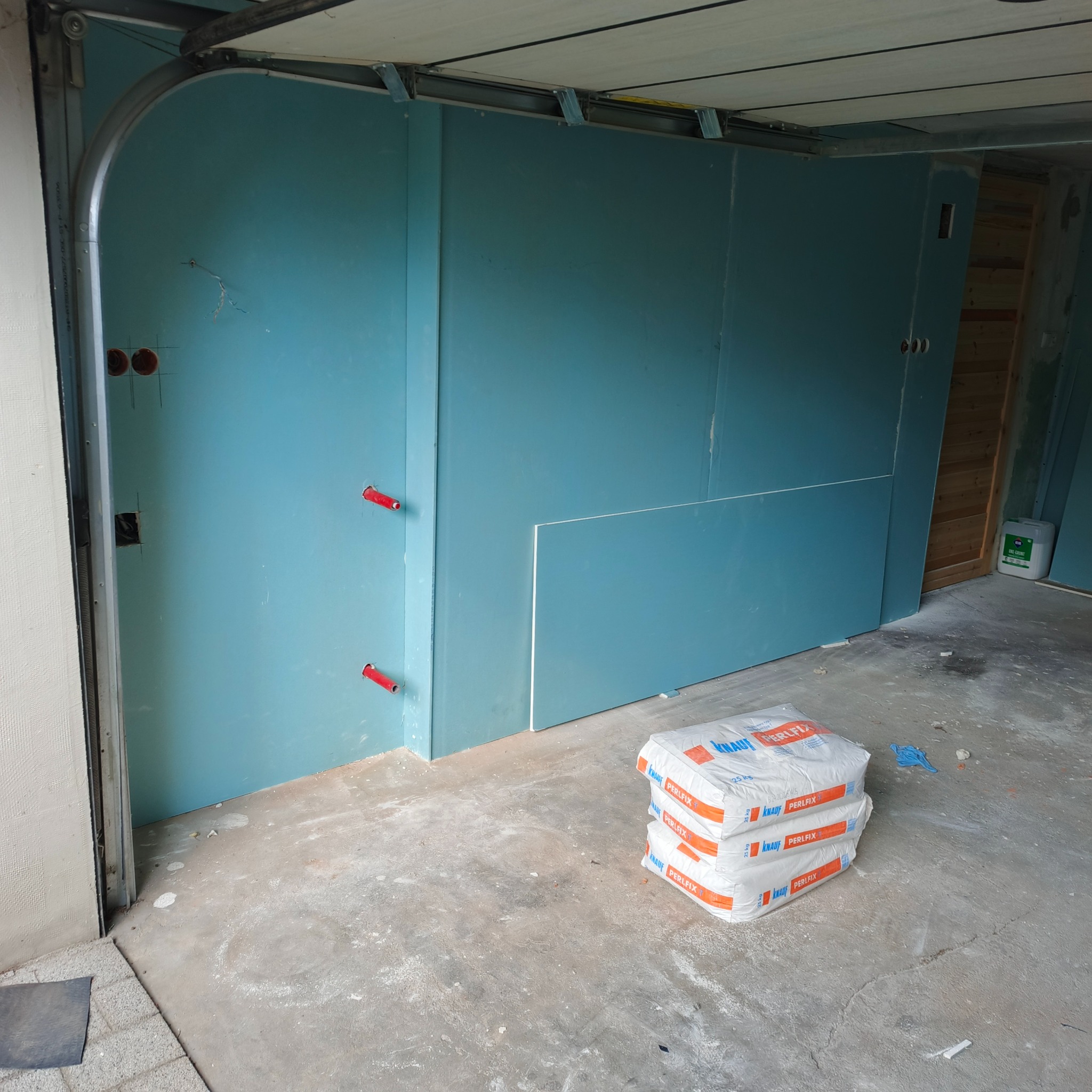 Montaż niebieskich płyt gipsowo-kartonowych na ścianie w garażu. Widoczne worki z zaprawą i fragment otwartej bramy garażowej. Etap prac wykończeniowych.