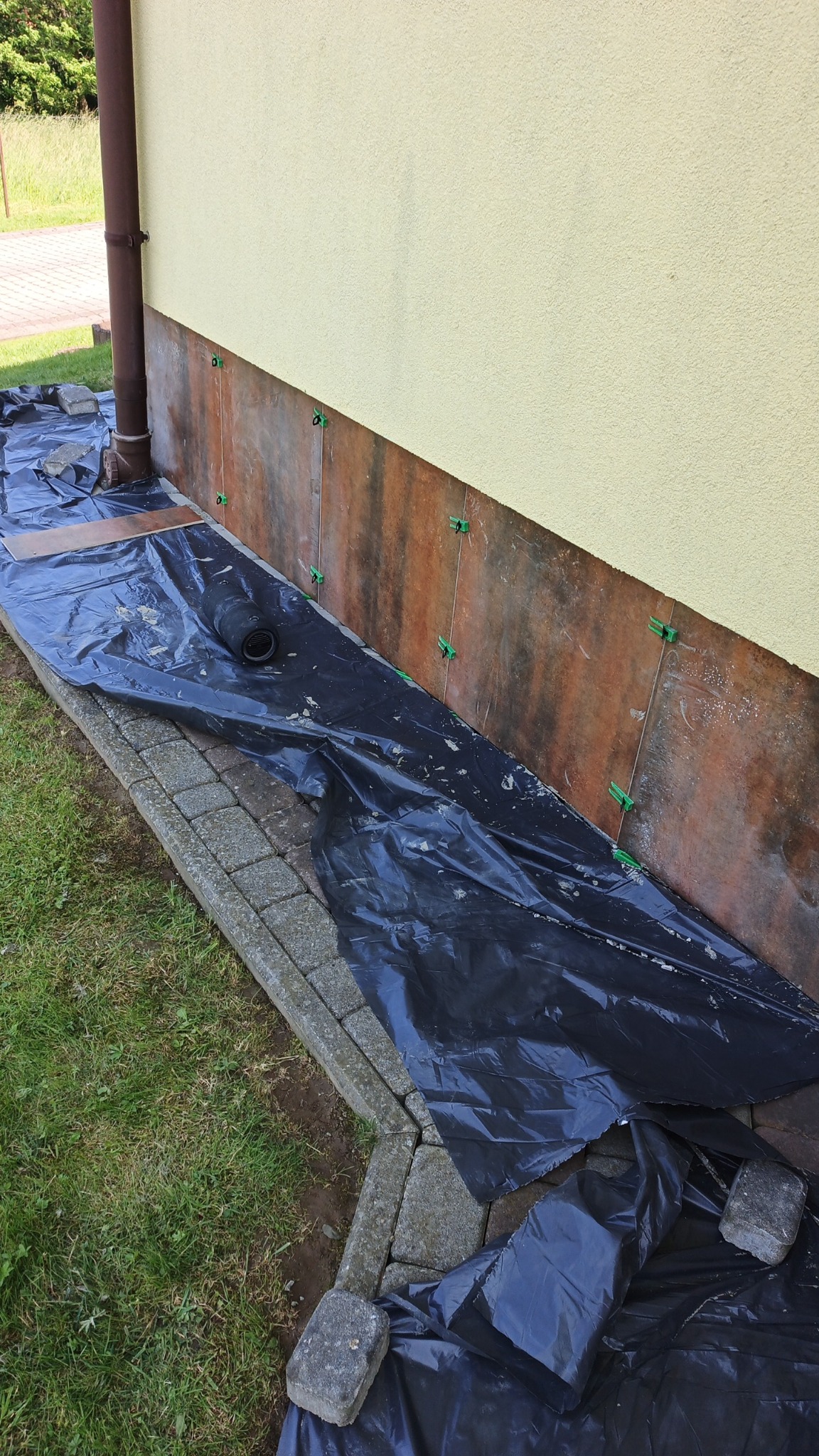 Montaż płytek elewacyjnych na cokole budynku z żółtą elewacją. Widoczne krzyżyki dystansowe i folia ochronna. Prace w toku, widok z zewnątrz.