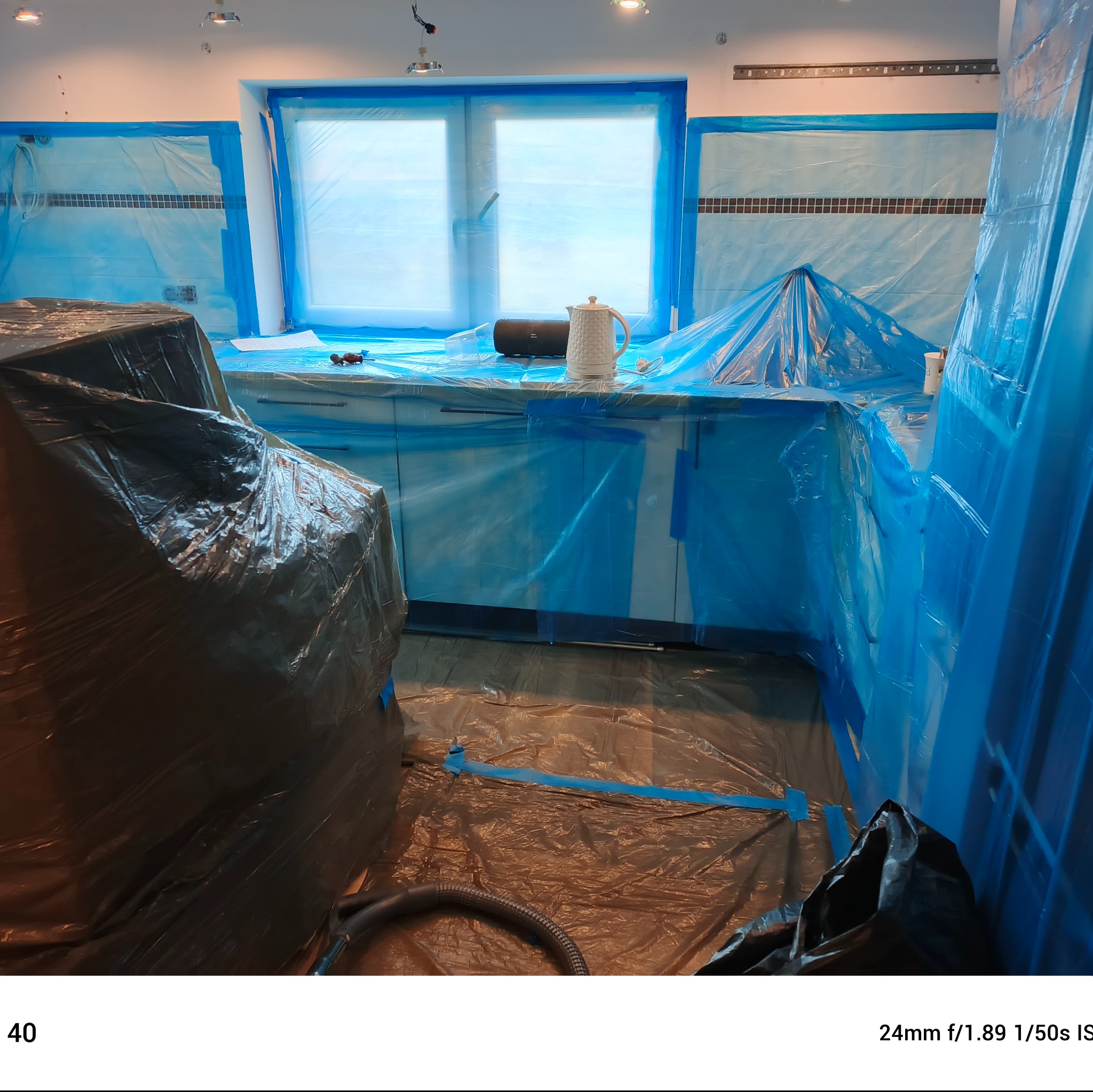 Kuchnia zabezpieczona folią malarską przed pracami. Widoczne okno, fragment blatu z czajnikiem i głośnikiem. Podłoga zakryta folią ochronną, wąż od odkurzacza.