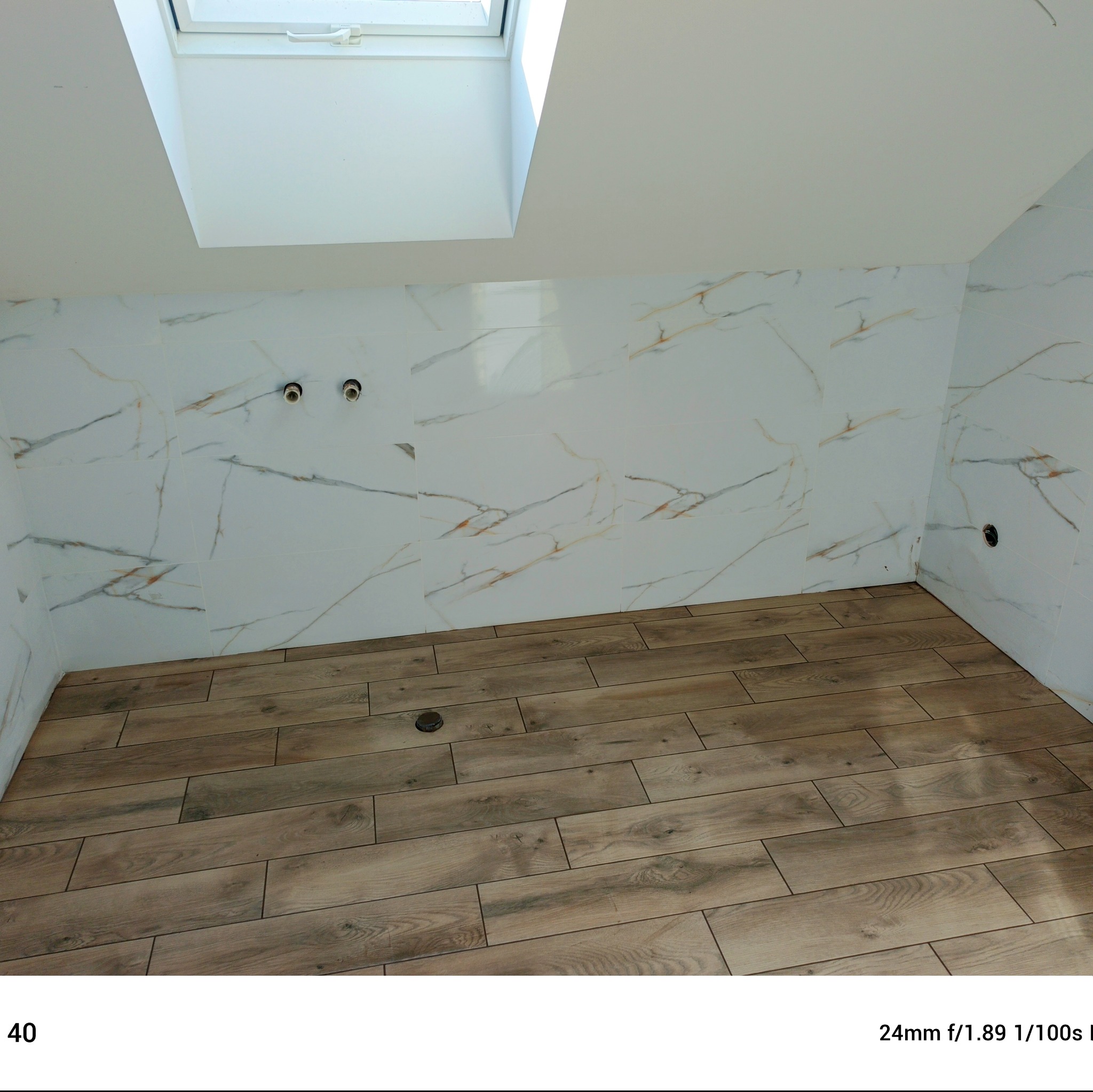 Jasna łazienka na poddaszu z drewnopodobną podłogą i marmurowymi płytkami ściennymi. Widoczne przyłącza wodne i okno dachowe.