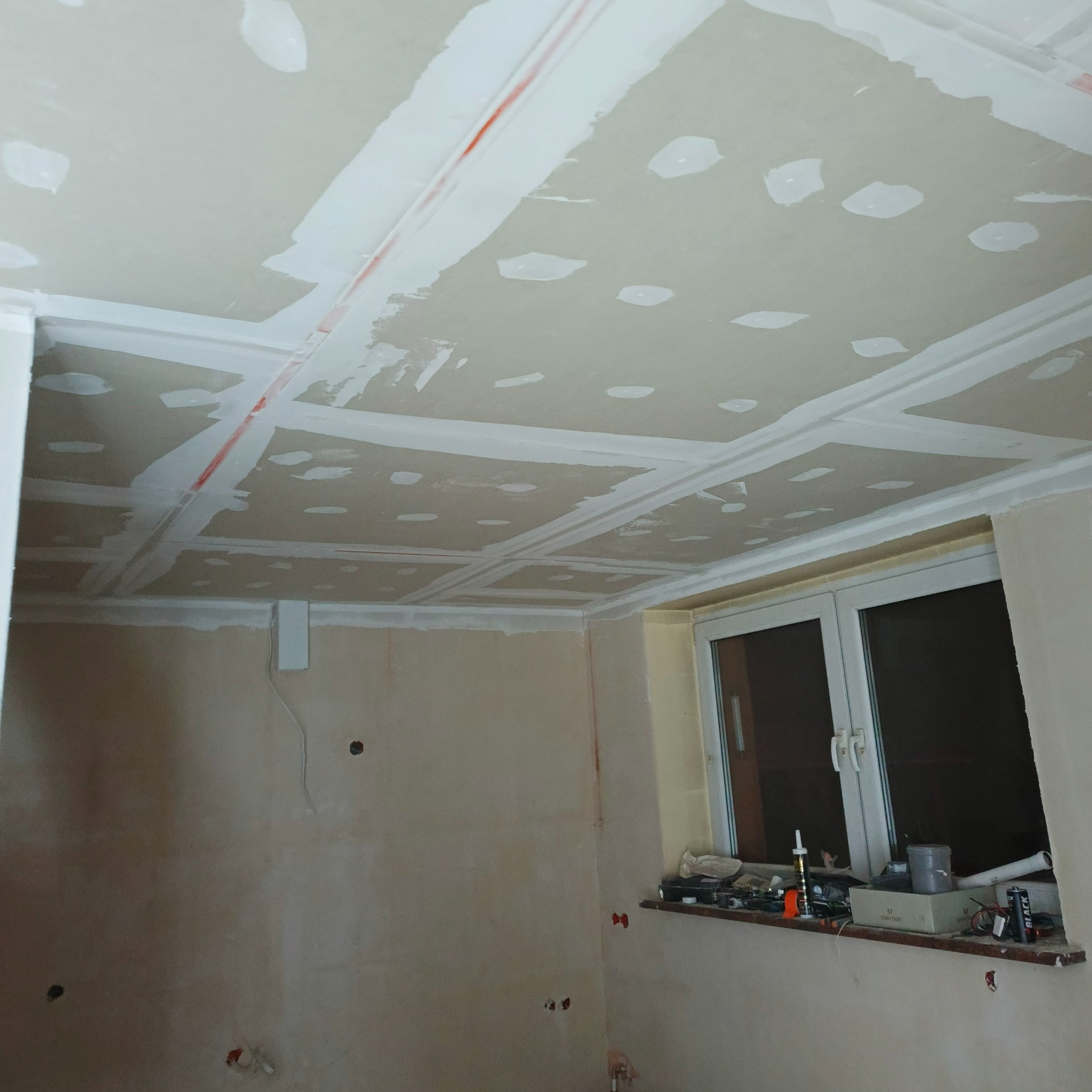 Sufit z gładzią gipsową w trakcie prac wykończeniowych, widoczne łączenia płyt kartonowo-gipsowych i fragment okna. Ściany przygotowane do malowania.