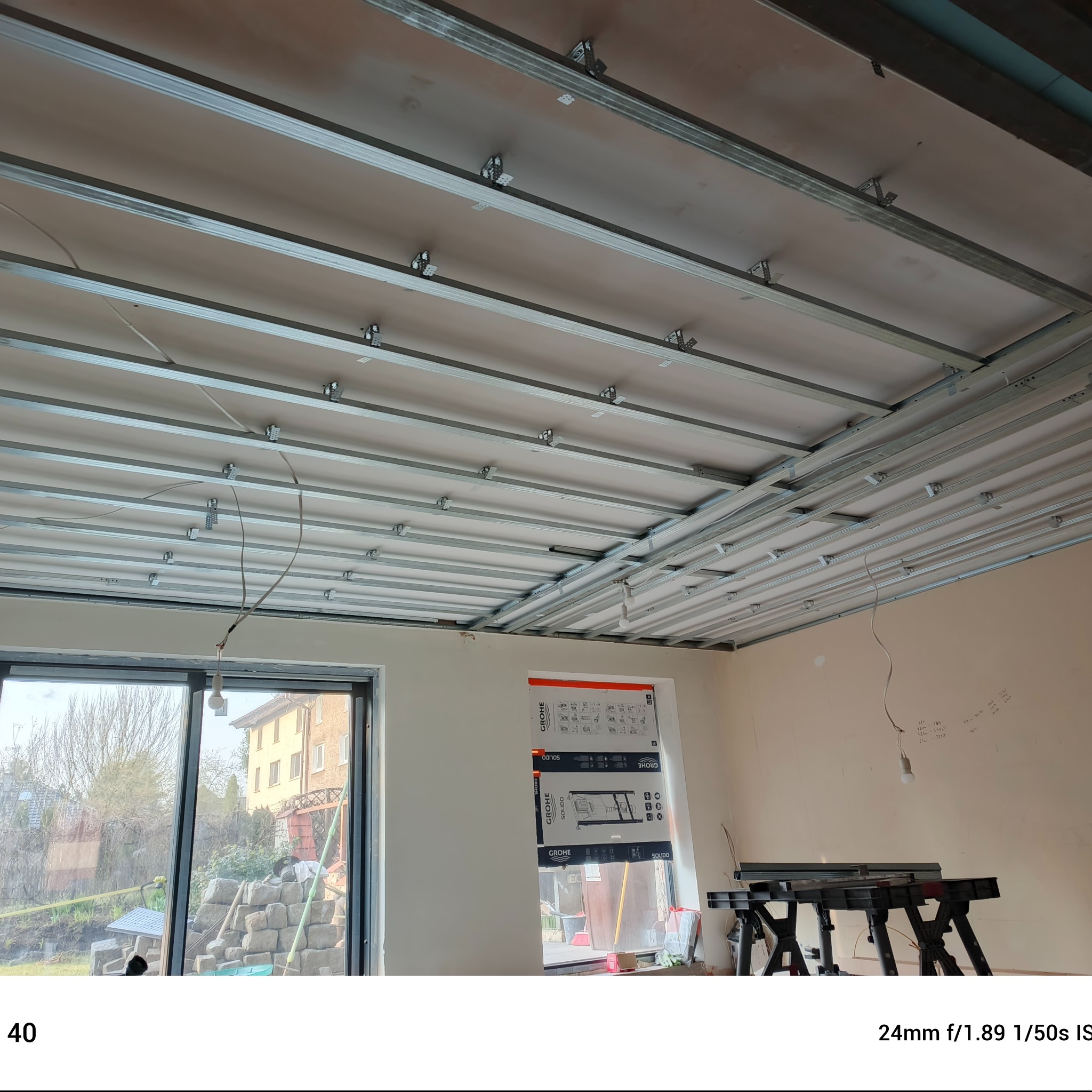 Montaż sufitu podwieszanego z metalową konstrukcją w jasnym wnętrzu. Widoczne profile, płyty gipsowe i narzędzia. W tle okno z widokiem na zieleń.