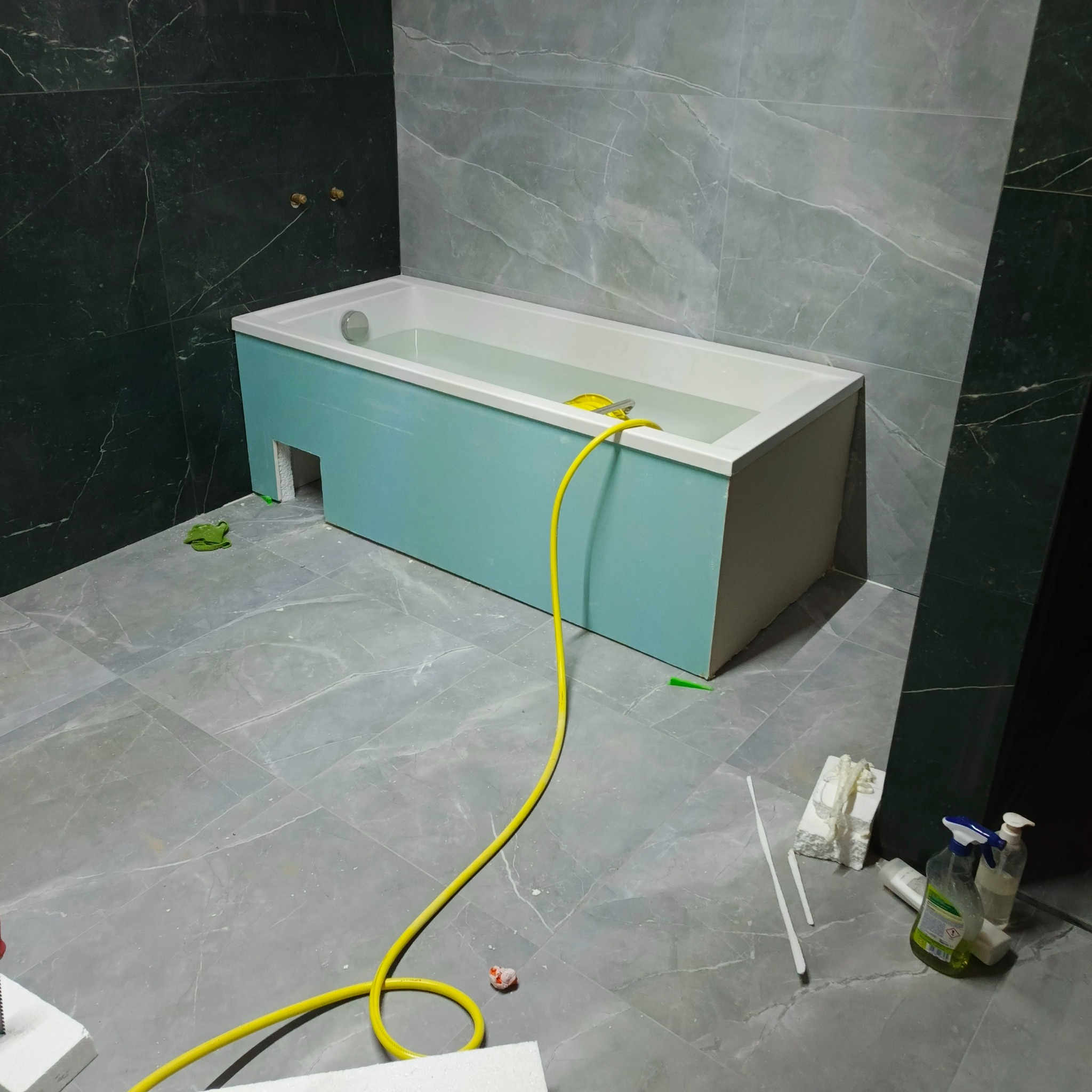 Narożna wanna z obudową w trakcie montażu w łazience wykończonej płytkami imitującymi marmur. Widoczne narzędzia i akcesoria instalacyjne.