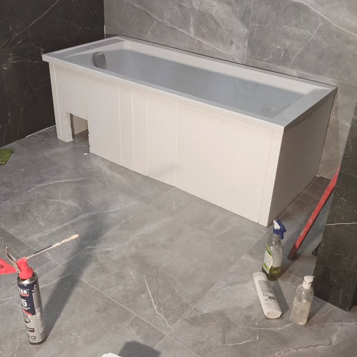 Biała wanna obudowana płytami w szarej łazience z marmurowymi płytkami. Na podłodze leżą narzędzia i środki czystości, co sugeruje etap prac wykończeniowych.