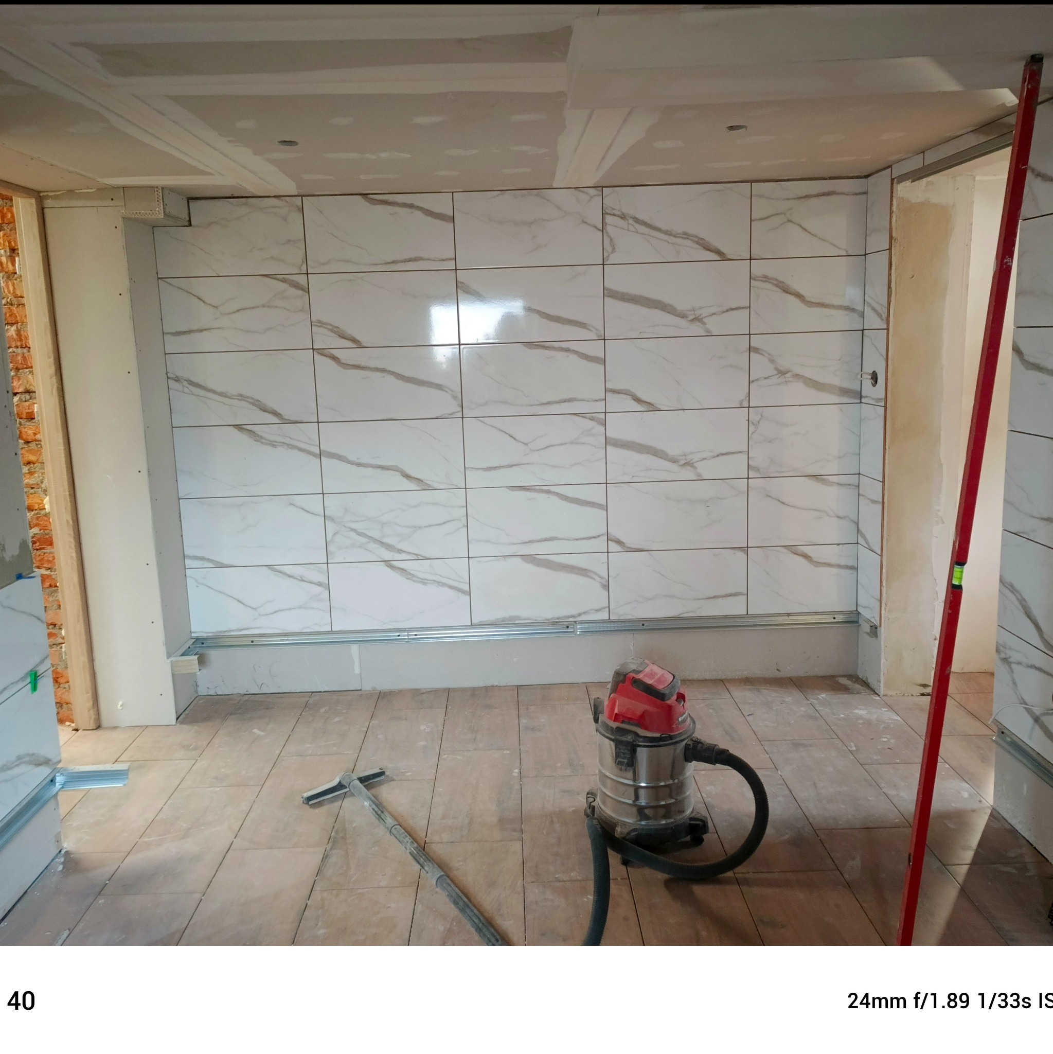 Wnętrze w trakcie prac: ściana z płytek imitujących marmur, widoczne profile montażowe, odkurzacz przemysłowy i poziomica. Etap prac wykończeniowych.