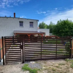 Nowoczesna, brązowa brama wjazdowa na posesję z domem w tle, widoczna furtka i poziome elementy konstrukcyjne, błękitne niebo.