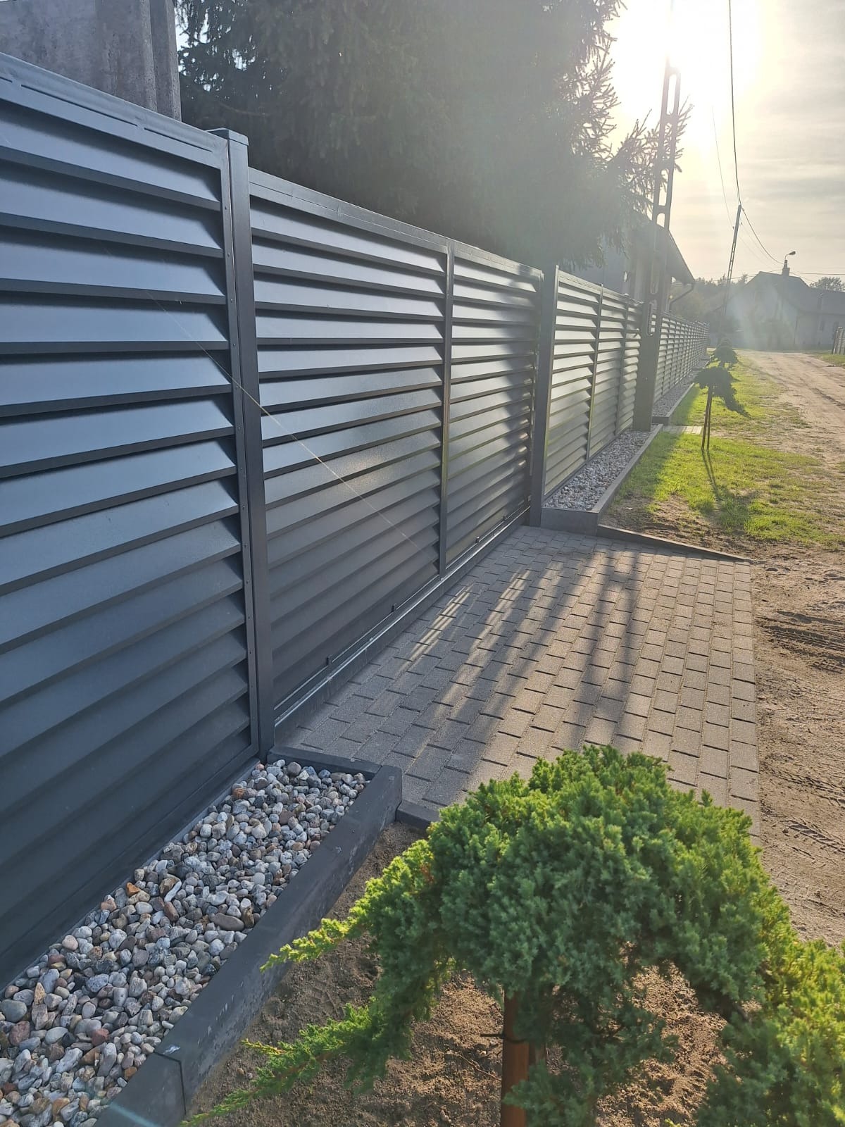 Nowoczesne, poziome, szare ogrodzenie lamelowe z podmurówką z kamienia, widok z perspektywy chodnika wyłożonego kostką brukową, z młodym iglakiem na pierwszym planie i domem w tle.