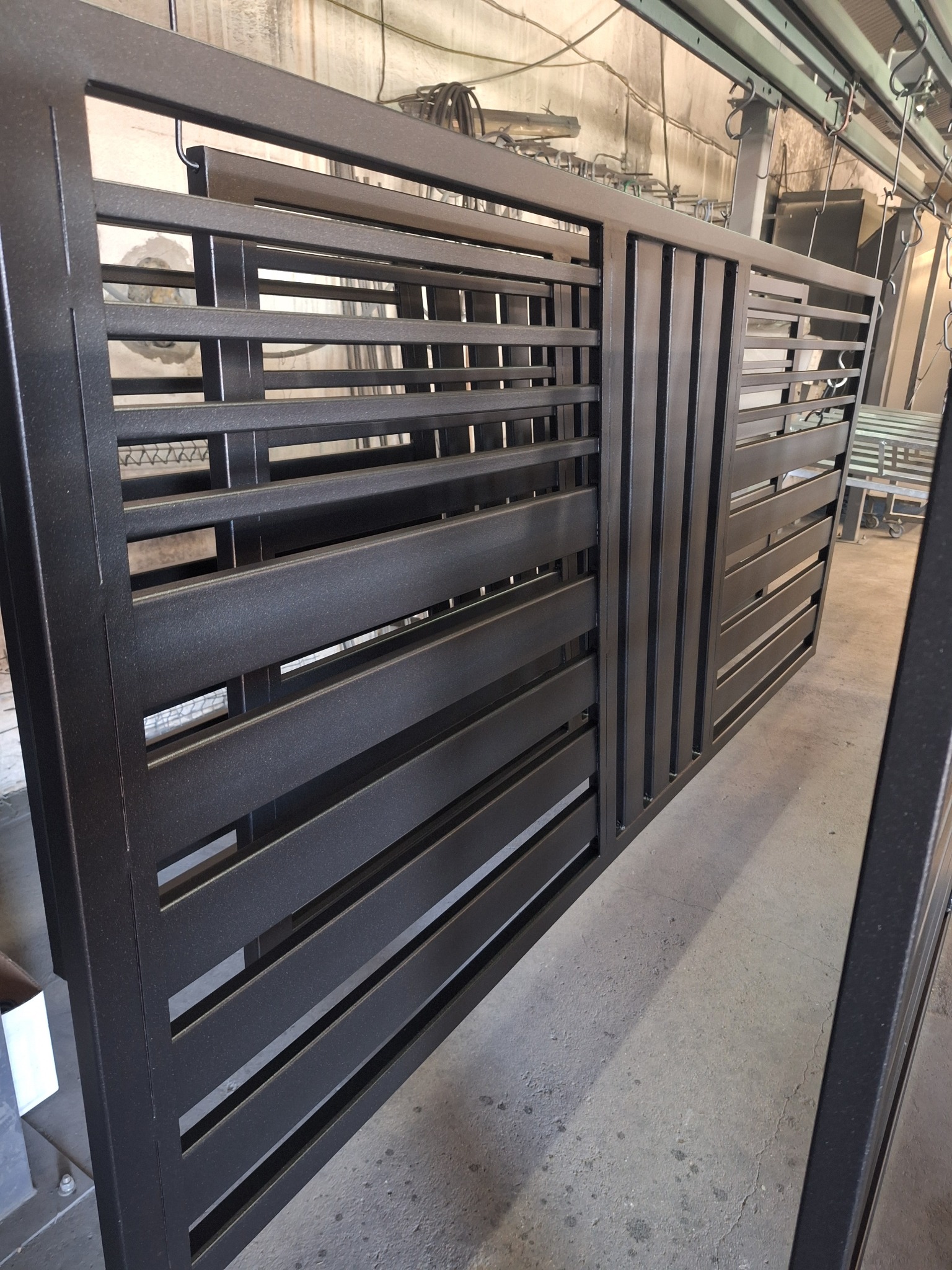 Metalowa brama wjazdowa z poziomymi szczeblami, świeżo pomalowana proszkowo na matowy czarny kolor, zawieszona na szynie w hali produkcyjnej.