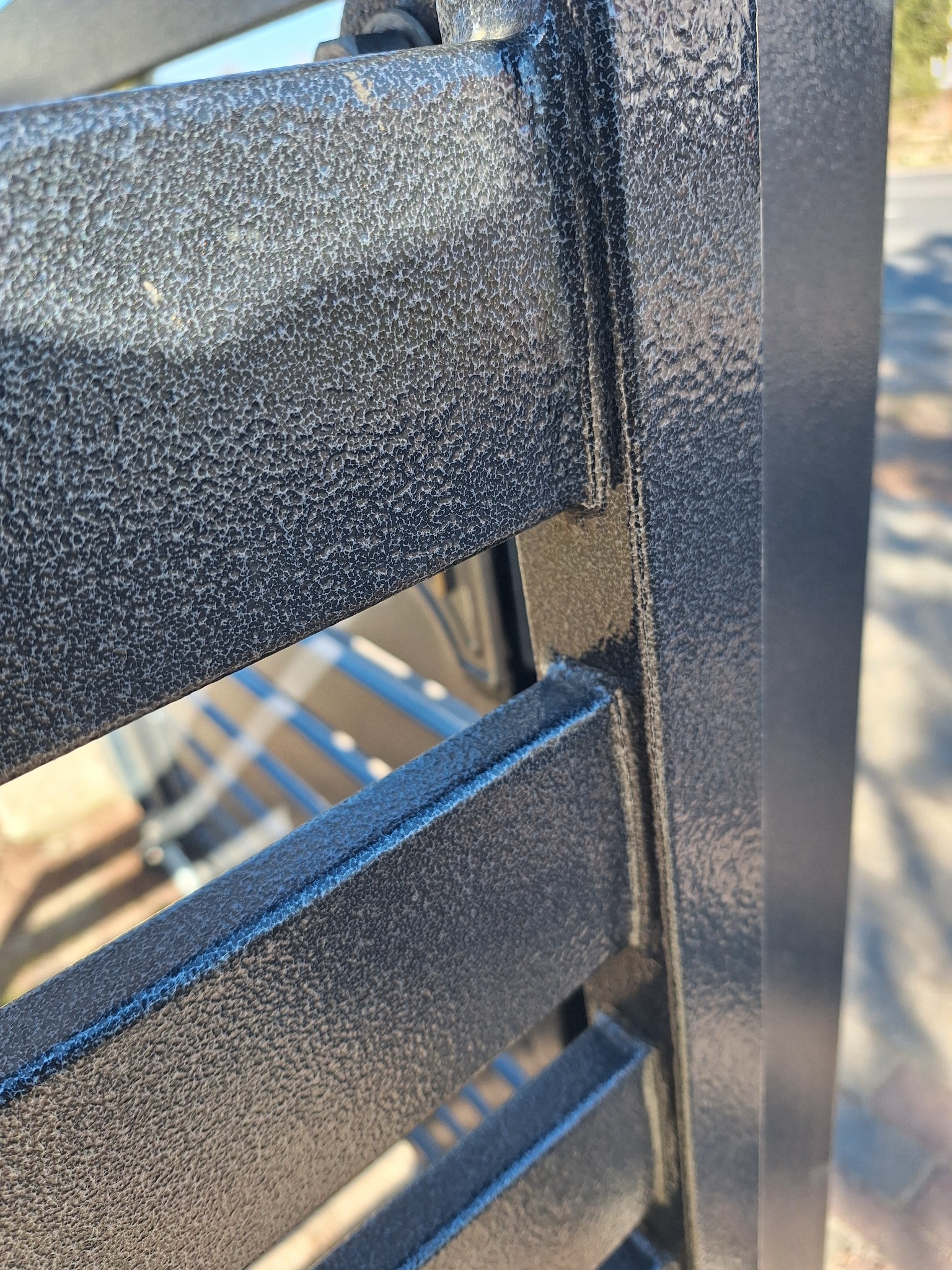 Fragment metalowej konstrukcji, prawdopodobnie ogrodzenia lub bramy, z wyraźną, teksturowaną powłoką w kolorze grafitowym, uchwycony w zbliżeniu, ukazujący detale łączeń i fakturę materiału.