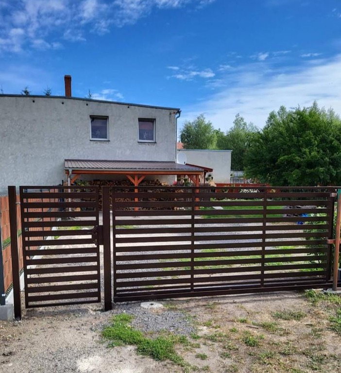 Nowoczesna, brązowa brama wjazdowa na posesję z domem w tle, widoczna furtka i poziome elementy konstrukcyjne, błękitne niebo.