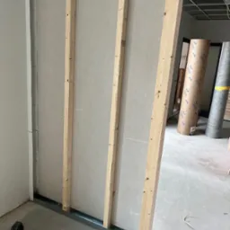 Konstrukcja ściany działowej z płyt kartonowo-gipsowych na metalowym stelażu, wzmocniona drewnianymi belkami, widoczny laserowy niwelator na podłodze.