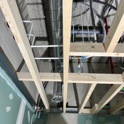 KRYSTIAN SASAL - Sufit podwieszany w trakcie montażu: widoczne drewniane belki konstrukcyjne, instalacja wentylacyjna z rurami spiralnymi, rury instalacji wodnej i elektrycznej, oraz fragment ściany z płyt...