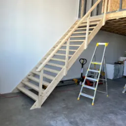 Surowe drewniane schody z barierką prowadzące na antresolę w pomieszczeniu w trakcie prac wykończeniowych, obok drabina aluminiowa i wózek paletowy.