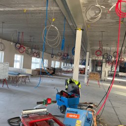 KRYSTIAN SASAL - Wnętrze w stanie surowym z widocznymi instalacjami elektrycznymi i hydraulicznymi zwisającymi z sufitu, narzędzia i wózek z urządzeniami na podłodze, widoczne okna w tle.