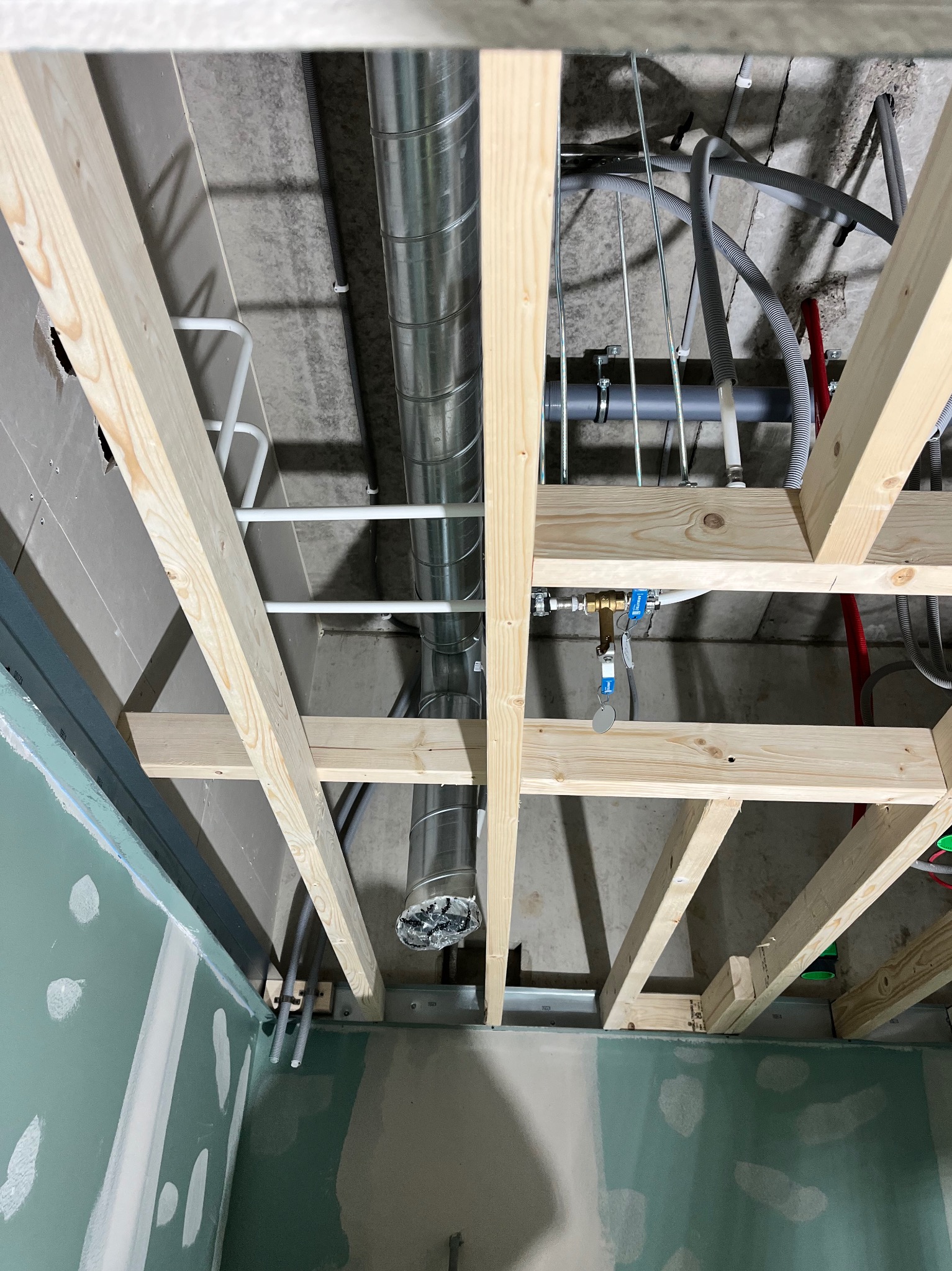 Sufit podwieszany w trakcie montażu: widoczne drewniane belki konstrukcyjne, instalacja wentylacyjna z rurami spiralnymi, rury instalacji wodnej i elektrycznej, oraz fragment ściany z płyt...