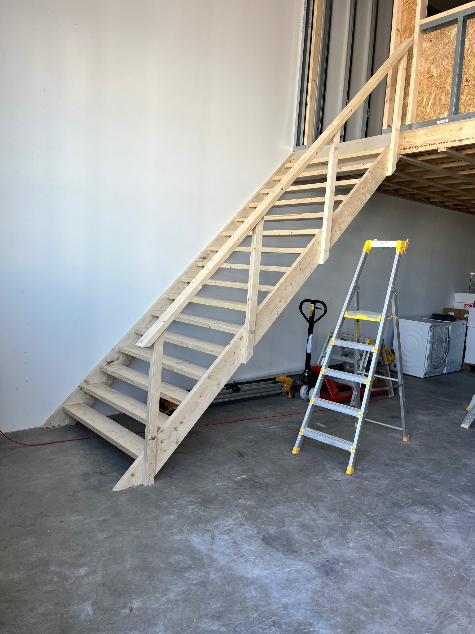 Surowe drewniane schody z barierką prowadzące na antresolę w pomieszczeniu w trakcie prac wykończeniowych, obok drabina aluminiowa i wózek paletowy.