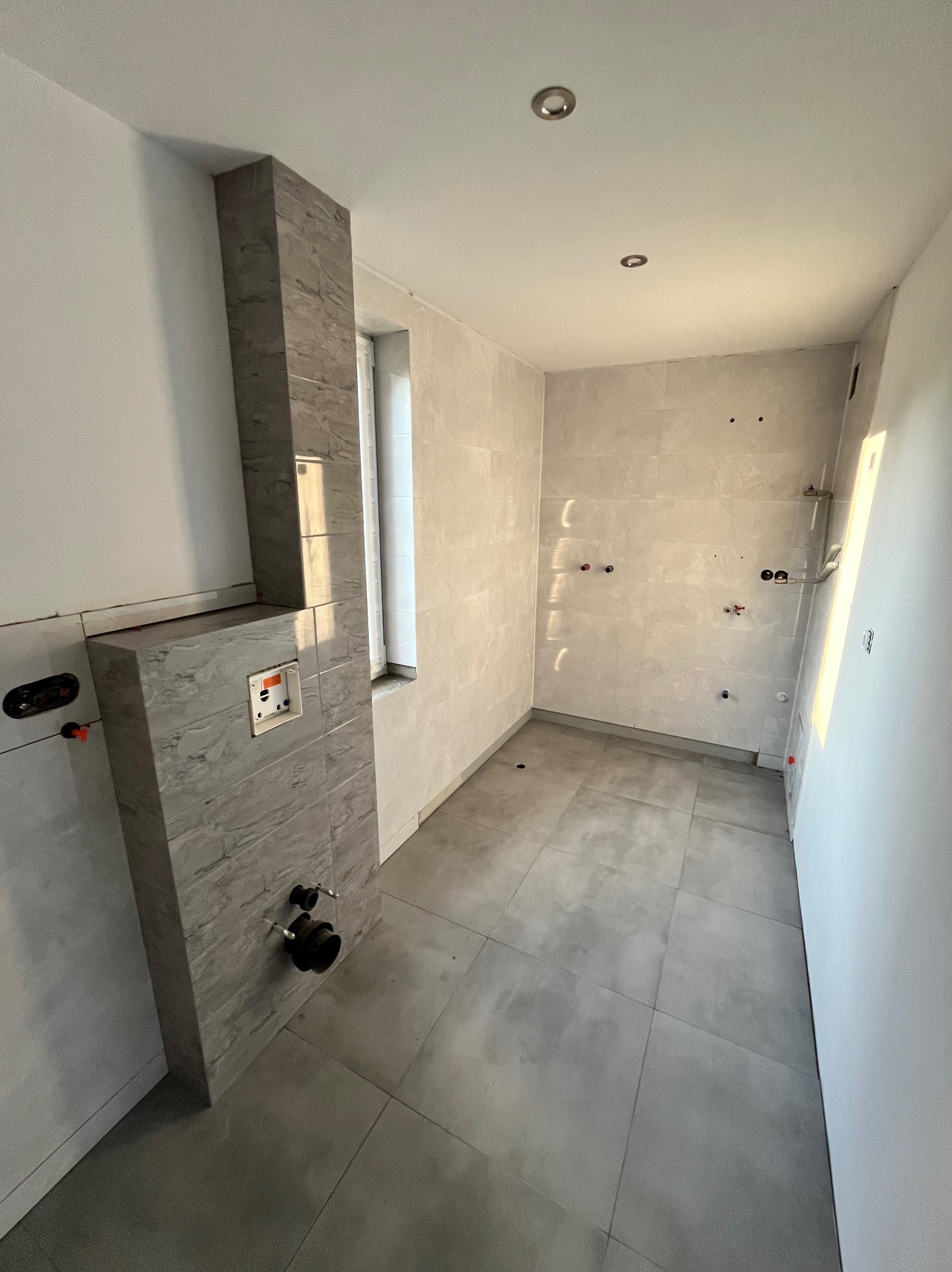 Wnętrze łazienki w trakcie prac wykończeniowych, widoczna zabudowa stelaża WC z szarych płytek imitujących marmur, surowe ściany z przygotowanymi punktami instalacji wodnej i elektrycznej,...