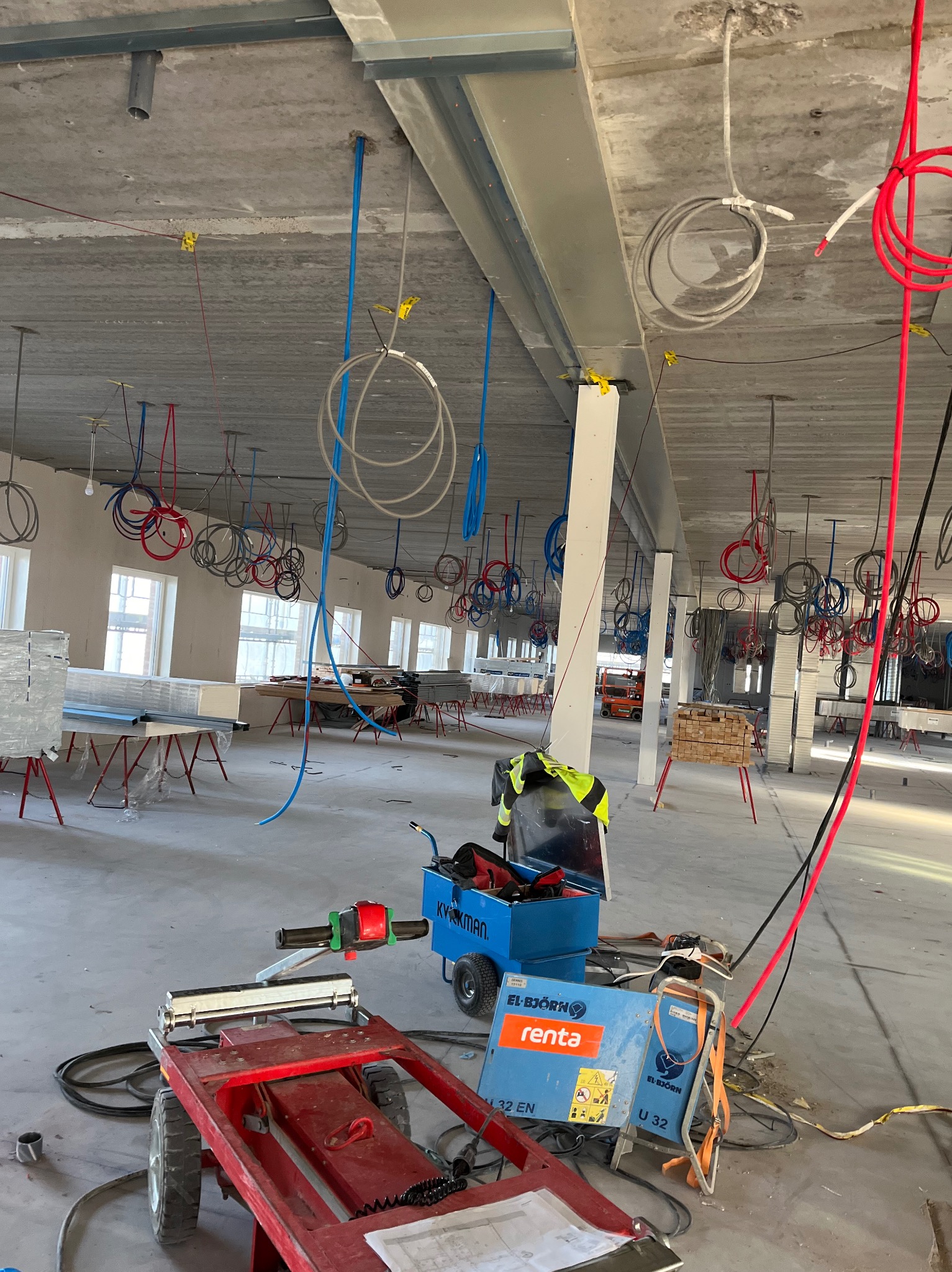 Wnętrze w stanie surowym z widocznymi instalacjami elektrycznymi i hydraulicznymi zwisającymi z sufitu, narzędzia i wózek z urządzeniami na podłodze, widoczne okna w tle.
