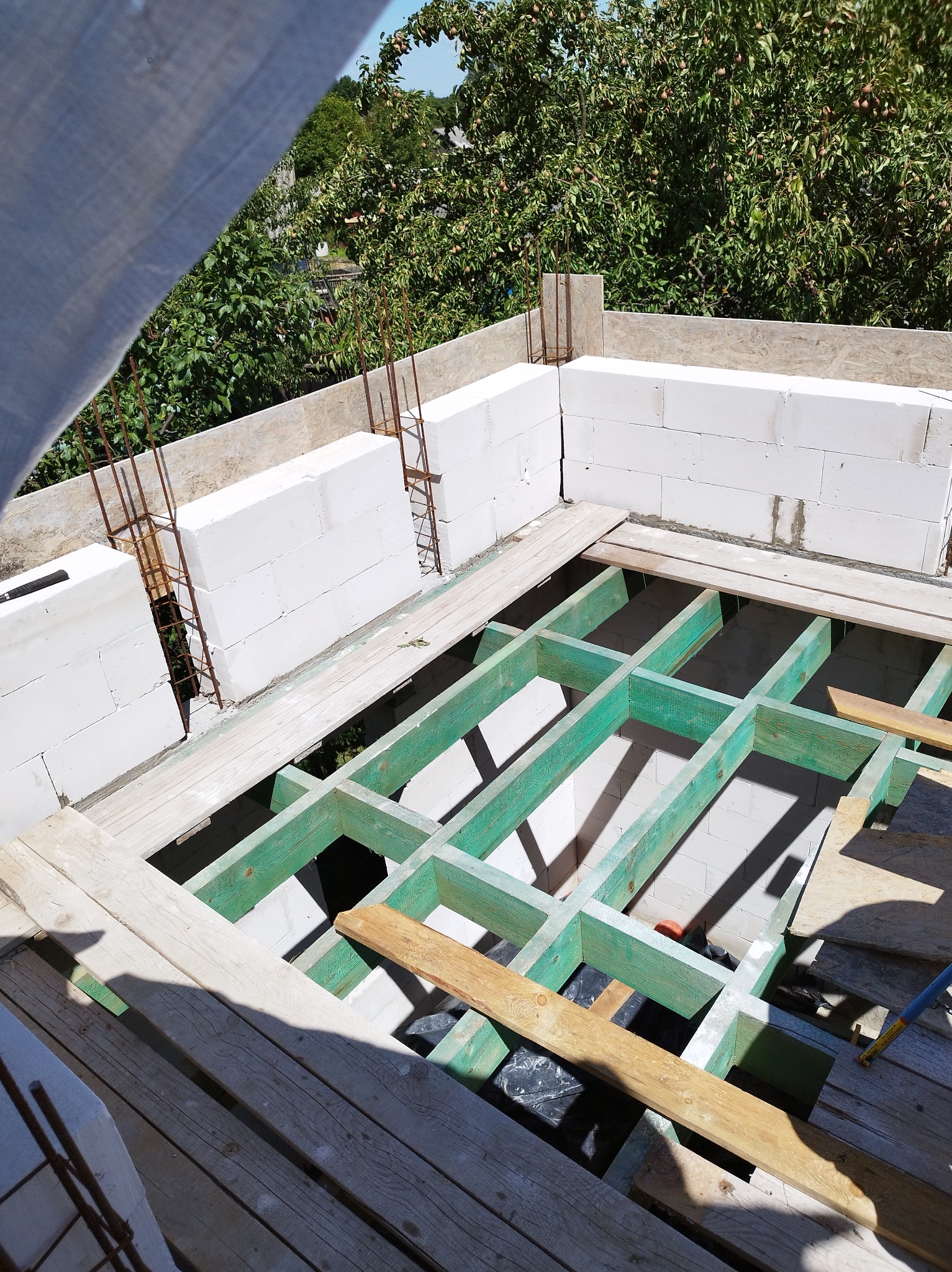 Wznoszenie ścian z białych bloczków betonu komórkowego Ytong na stropie z zielonej drewnianej konstrukcji, widoczne zbrojenie i drzewa w tle.