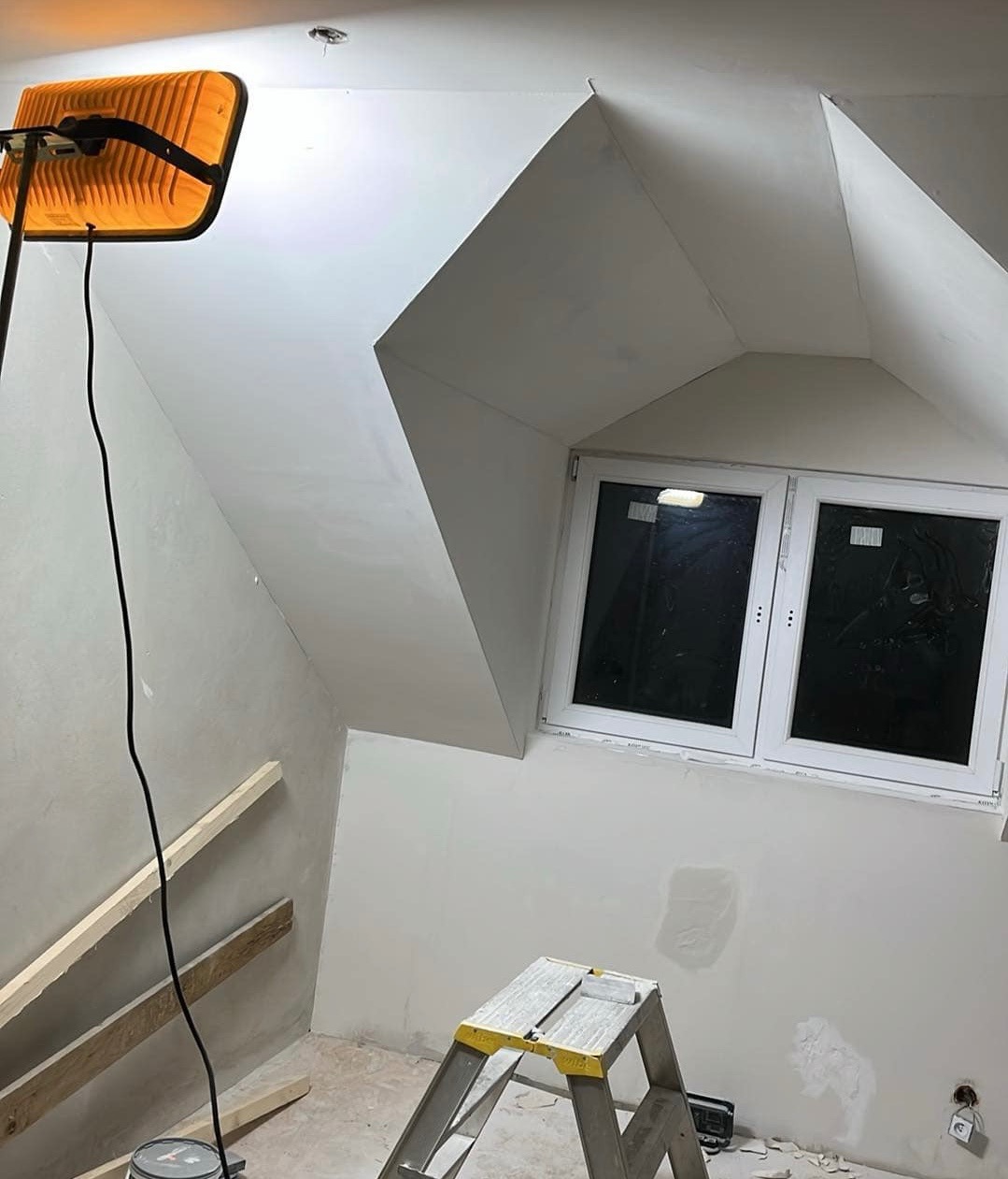 Poddasze w trakcie prac wykończeniowych: ściany po szpachlowaniu, oświetlone lampą halogenową, widoczna drabina, okno PCV, fragment drewnianej konstrukcji i ślady po pracach na podłodze.