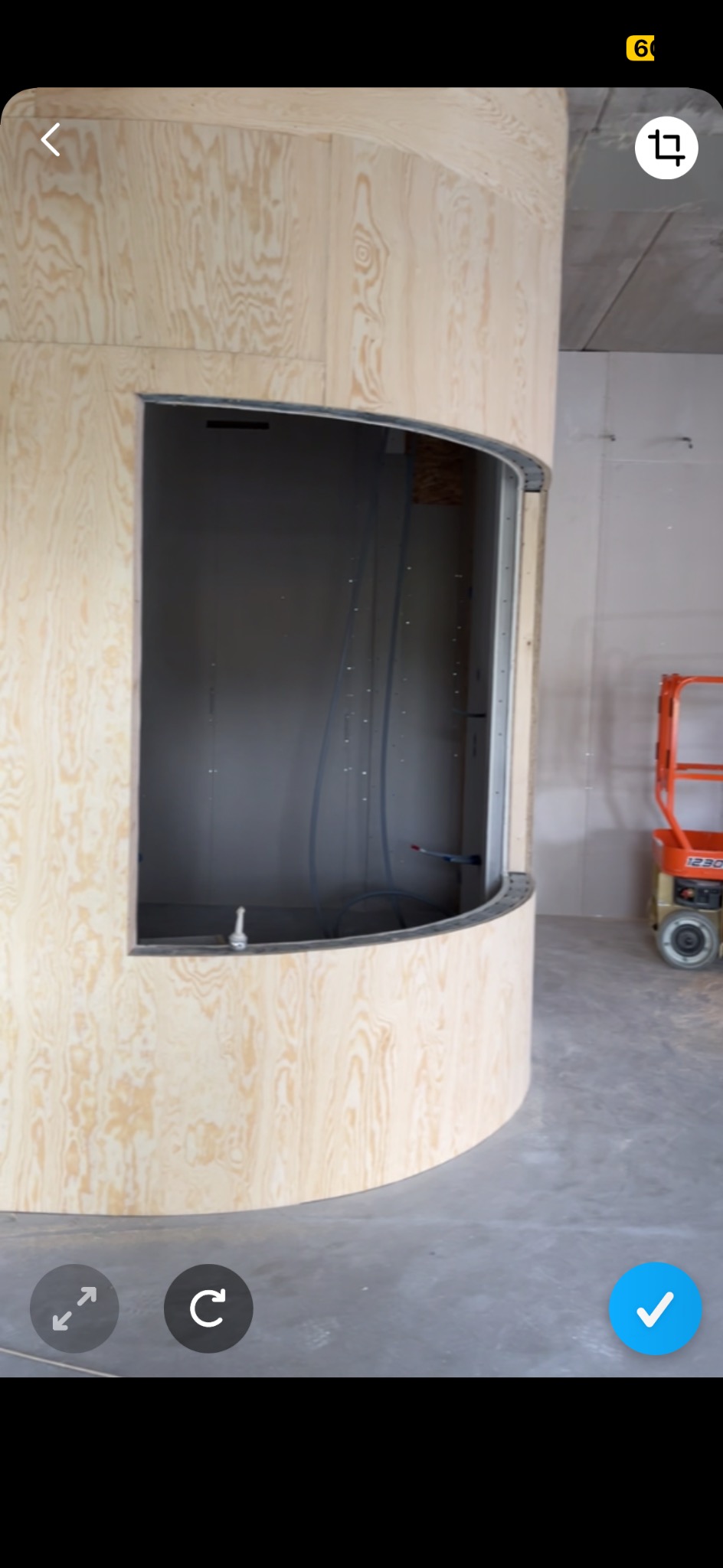 Konstrukcja z surowej sklejki tworząca zaokrąglony element architektoniczny z otworem, widoczne instalacje wewnątrz, w tle podnośnik nożycowy na budowie.