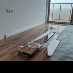 Rozpakowane panele podłogowe Wenninger w trakcie układania, widoczne fragmenty ułożonej podłogi z połyskiem i listwy przypodłogowe, w tle okno balkonowe z widokiem na zamglony krajobraz.