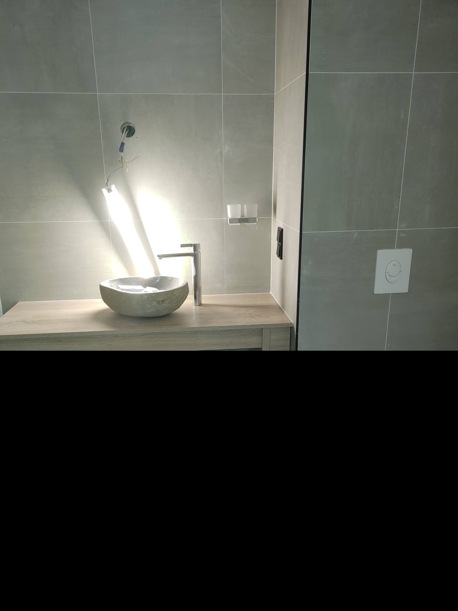 Umywalka w kształcie misy z kamienia, zamontowana na drewnianym blacie w łazience z szarymi płytkami, widoczne podłączenia elektryczne i przycisk spłuczki.