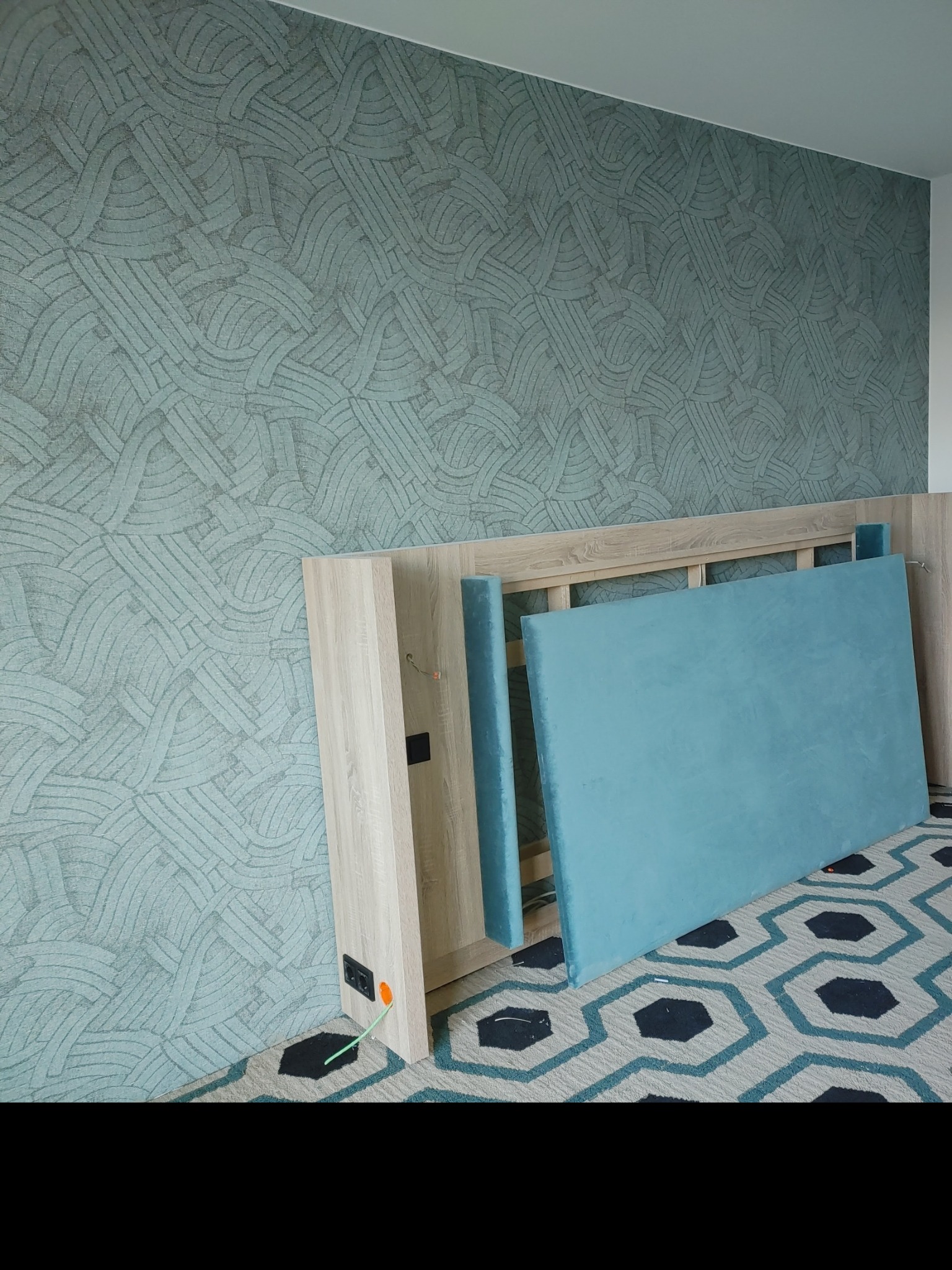 Ściana pokryta tapetą o abstrakcyjnym wzorze w odcieniach szarości i błękitu, z fragmentami rozłożonego łóżka z jasnego drewna i turkusowym zagłówkiem.