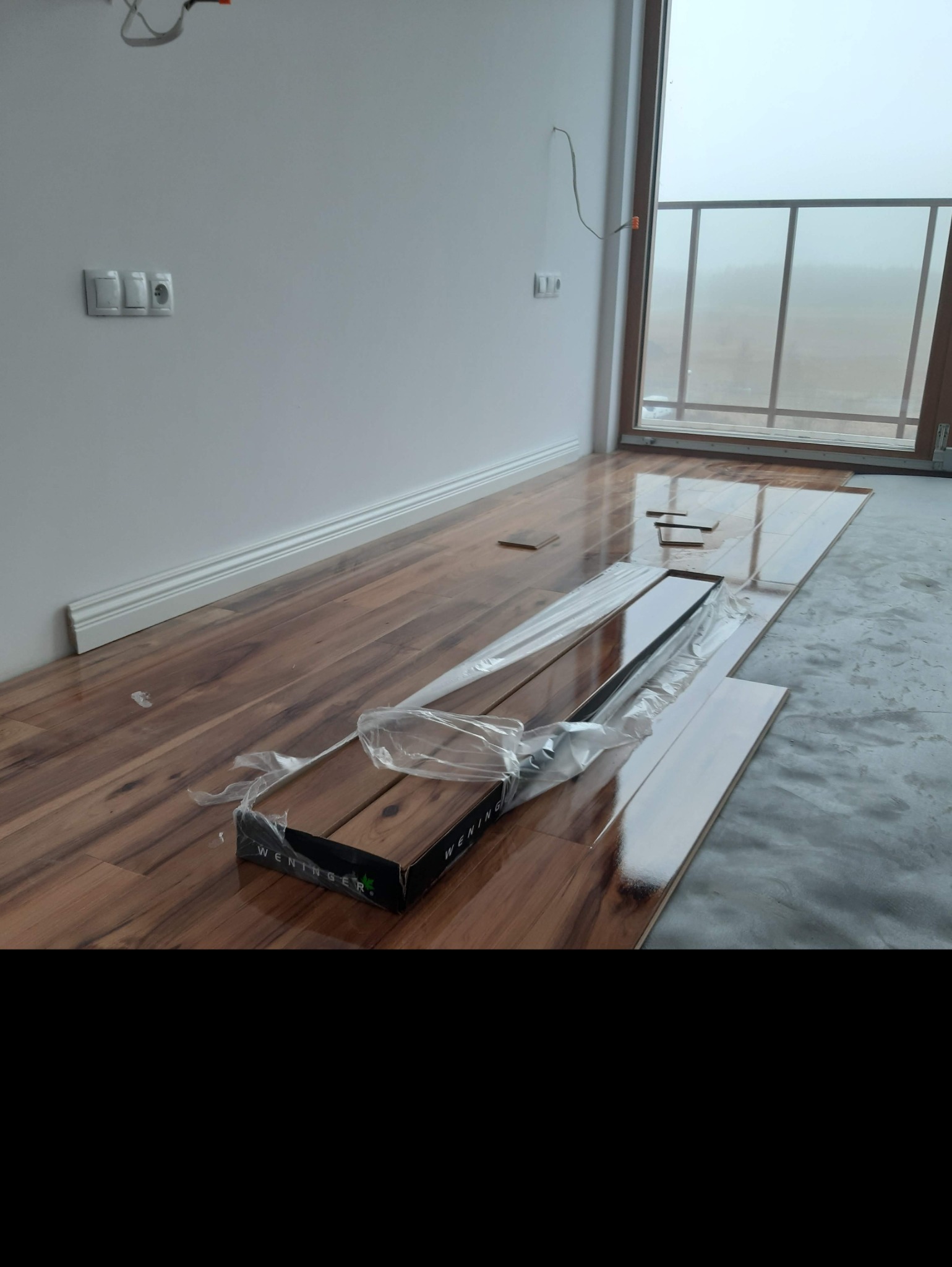 Rozpakowane panele podłogowe Wenninger w trakcie układania, widoczne fragmenty ułożonej podłogi z połyskiem i listwy przypodłogowe, w tle okno balkonowe z widokiem na zamglony krajobraz.
