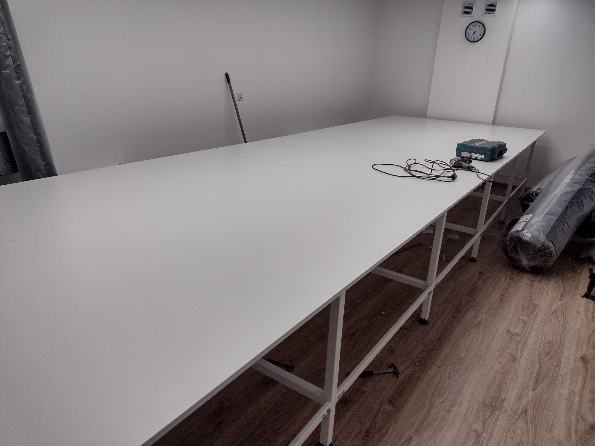 Długa, biała szafa na wymiar z narzędziami na blacie w jasnym pomieszczeniu. Widoczny zegar na ścianie i zwinięty materiał w tle. Minimalistyczny design.