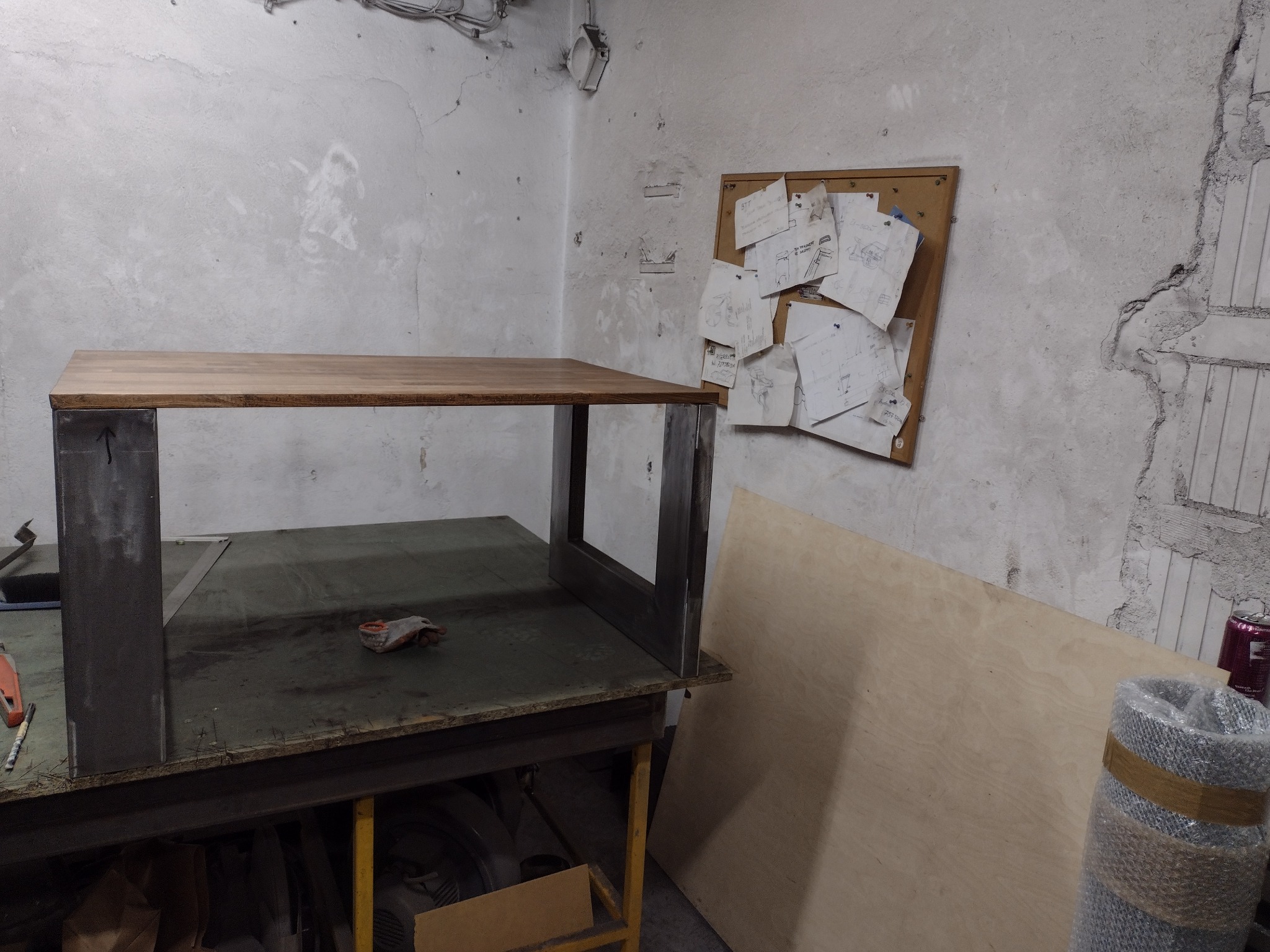 Stół z drewnianym blatem i metalową ramą w warsztacie. Na ścianie tablica z projektami. Widoczne narzędzia i materiały. Surowe wnętrze.