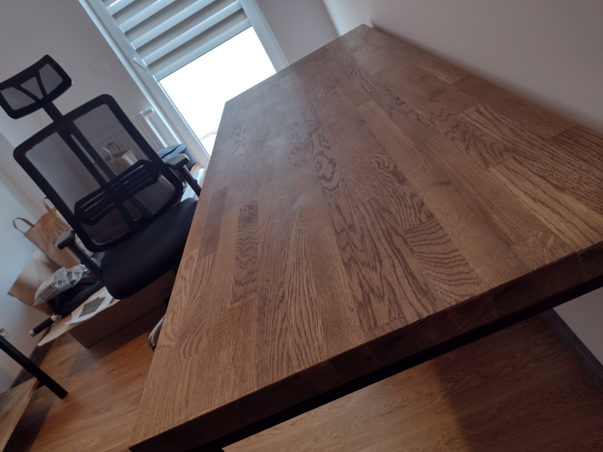 Drewniany blat biurka o wyraźnym usłojeniu, ustawiony pod kątem w jasnym pomieszczeniu. W tle widoczne krzesło biurowe i okno z żaluzjami.