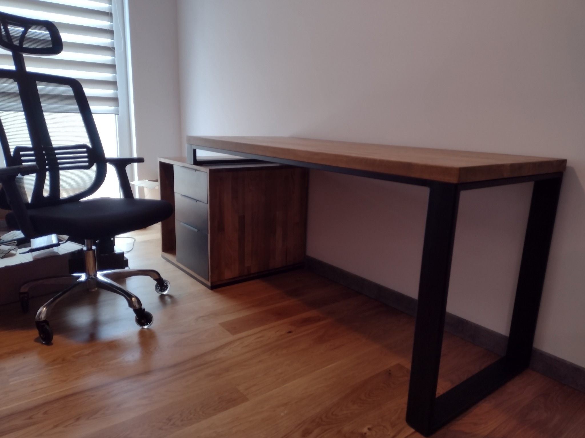 Nowoczesne biurko z drewnianym blatem i czarnymi metalowymi nogami, obok krzesło biurowe z siatkowym oparciem na drewnianej podłodze. Minimalistyczny design.