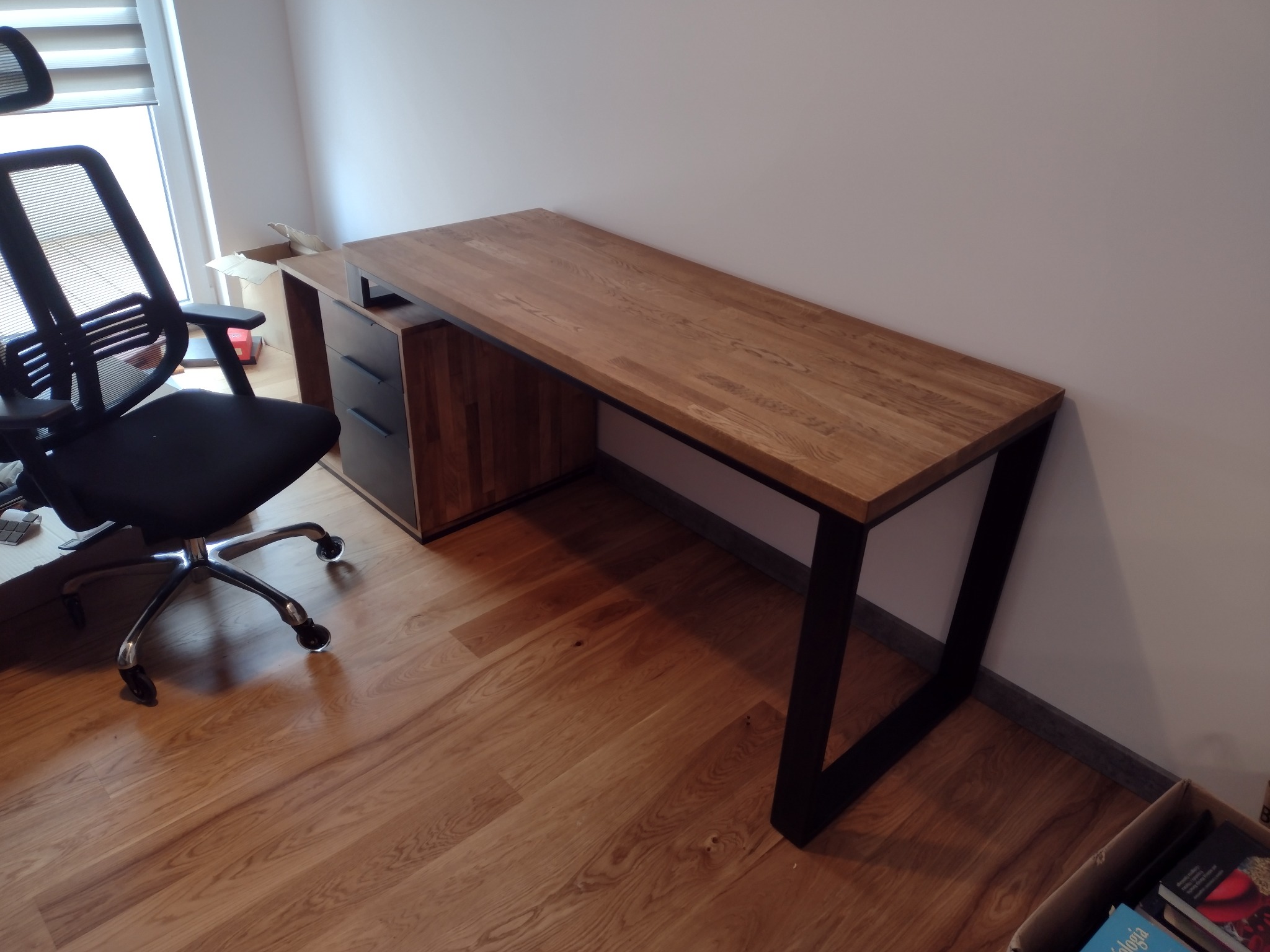 Dębowe biurko na metalowych nogach, zintegrowane z szafką z szufladami, stoi na drewnianej podłodze obok czarnego krzesła biurowego.