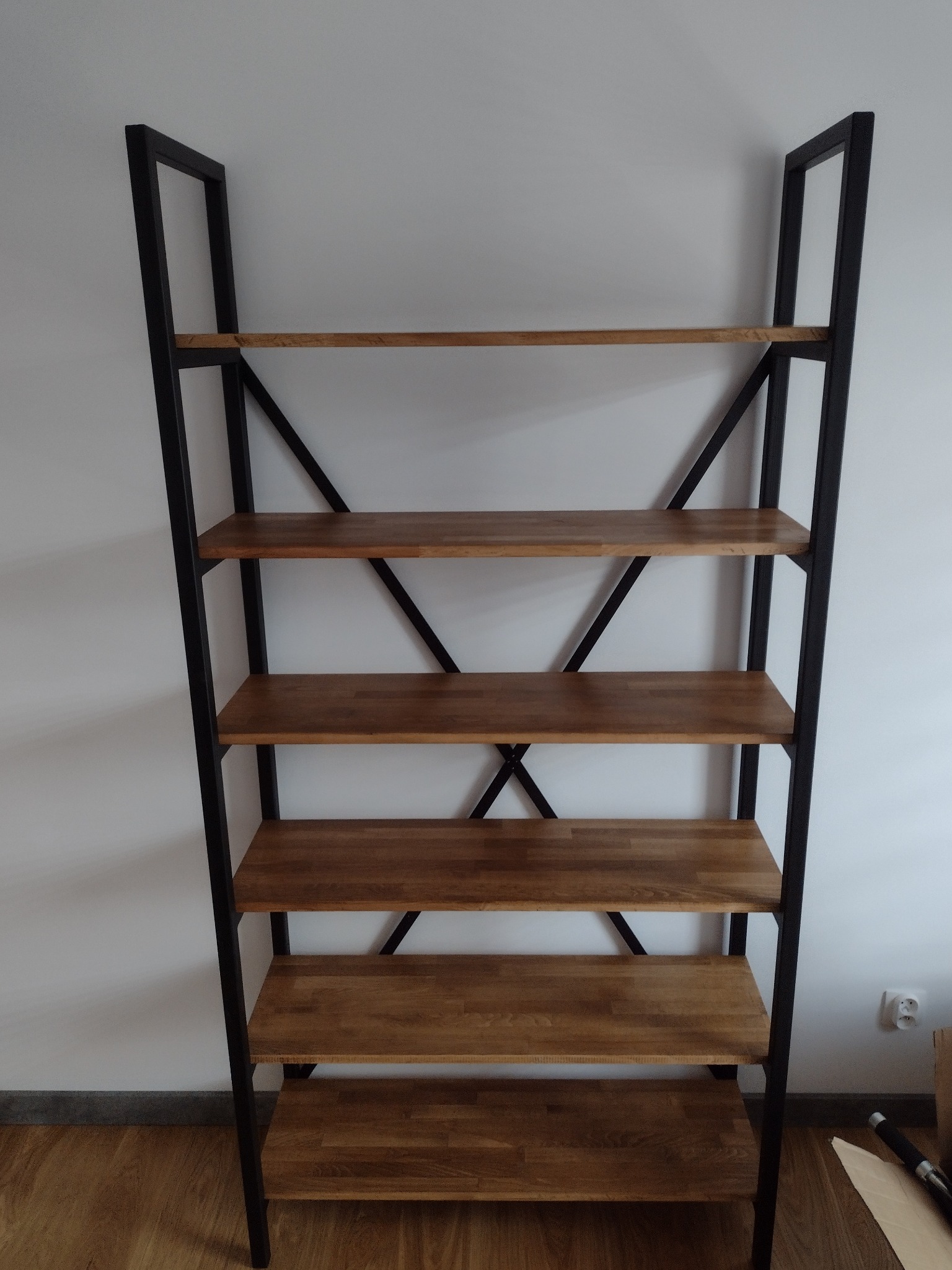 Regał w stylu loftowym z drewnianymi półkami i czarną, metalową konstrukcją, ustawiony na podłodze przy białej ścianie. Widoczny fragment gniazdka elektrycznego.