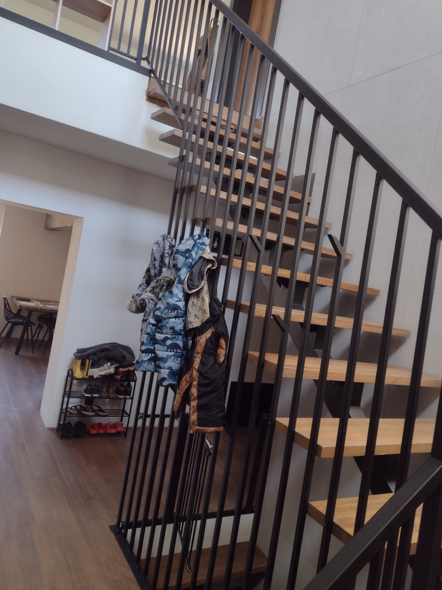 Nowoczesne schody z drewnianymi stopniami i czarną metalową balustradą, widoczne z dołu, z elementami garderoby na balustradzie i półką na buty w tle.
