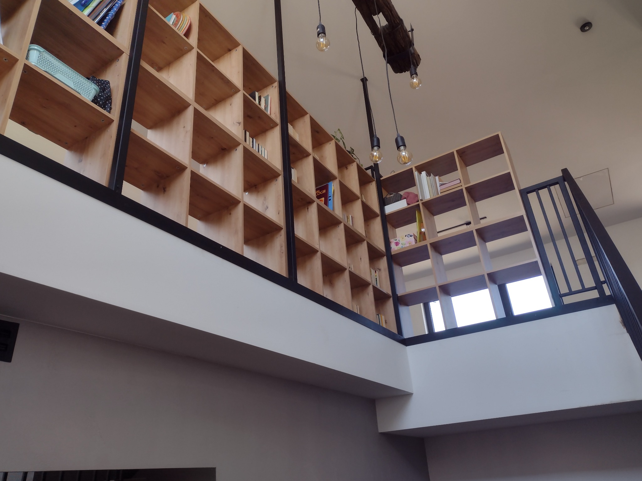 Dwupoziomowa, drewniana biblioteczka z książkami i dekoracjami, oświetlona wiszącymi lampami, widziana z dołu w jasnym wnętrzu.