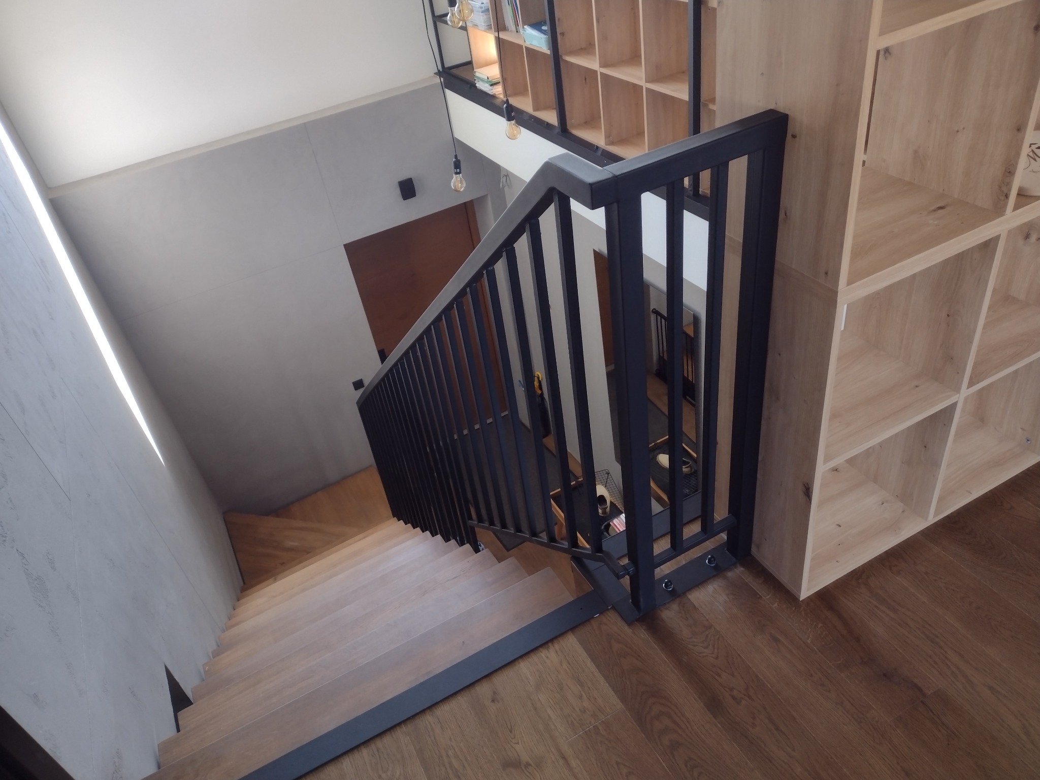 Nowoczesne schody drewniane z czarną metalową balustradą. Widok z góry na stopnie i poręcze. W tle drewniany regał z półkami i szara ściana.