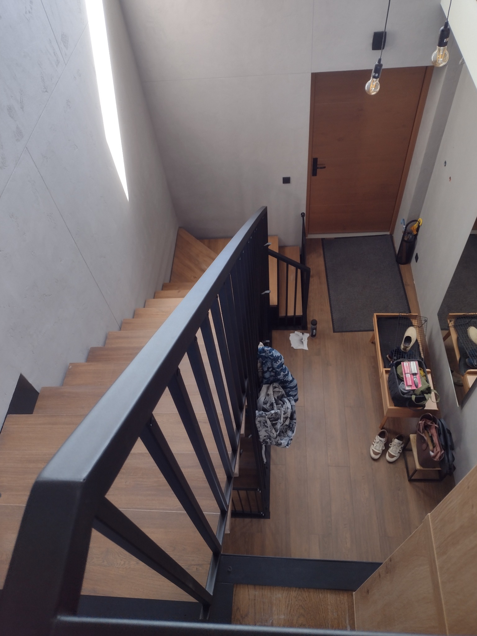 Minimalistyczne schody z drewnianymi stopniami i czarną, metalową balustradą, widziane z góry. W tle drzwi wejściowe i elementy wyposażenia wnętrza.