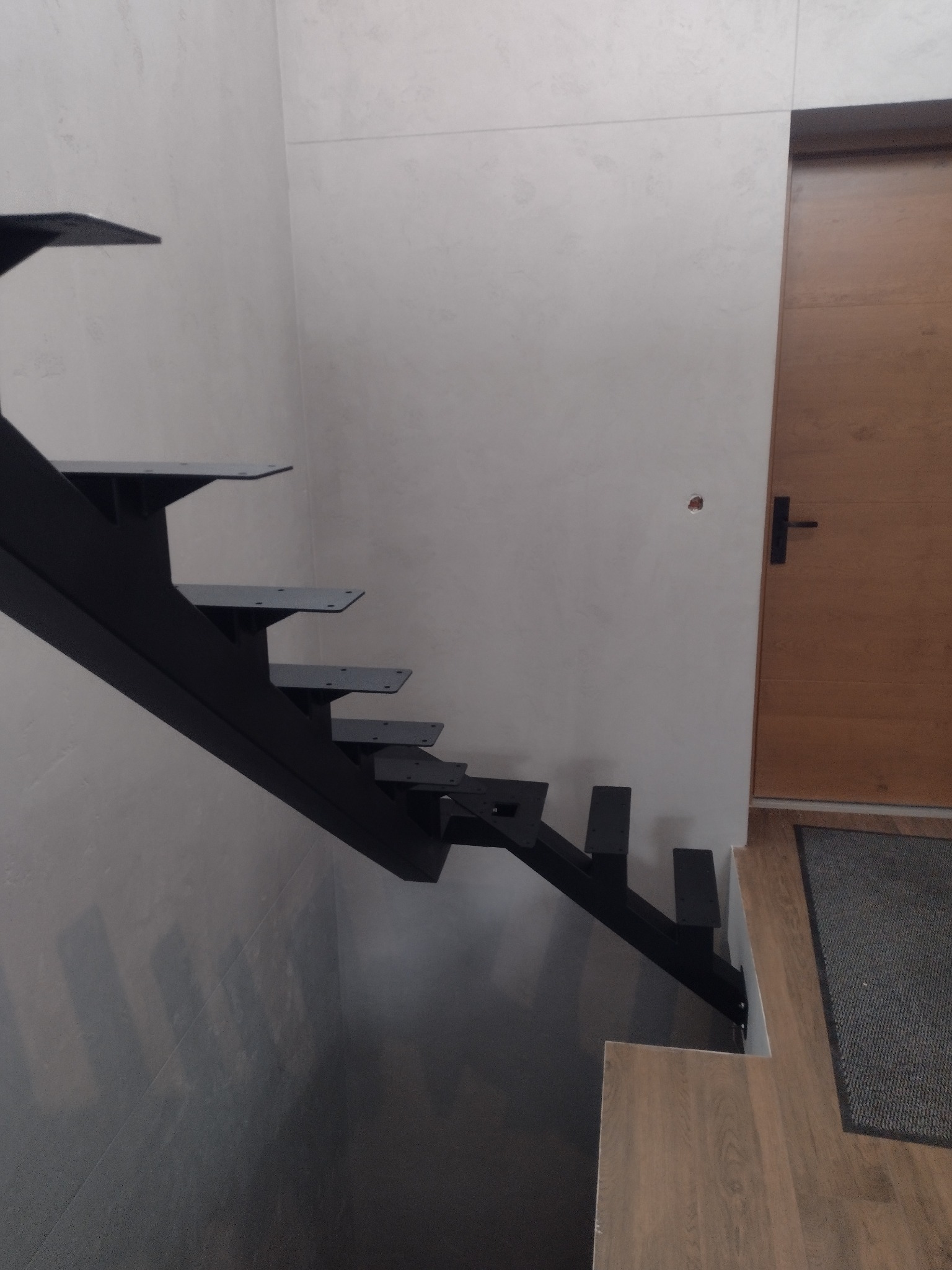 Minimalistyczne, czarne schody metalowe wspornikowe, z widocznymi mocowaniami do ściany, na tle jasnoszarej ściany i drewnianych drzwi, nowoczesny design.