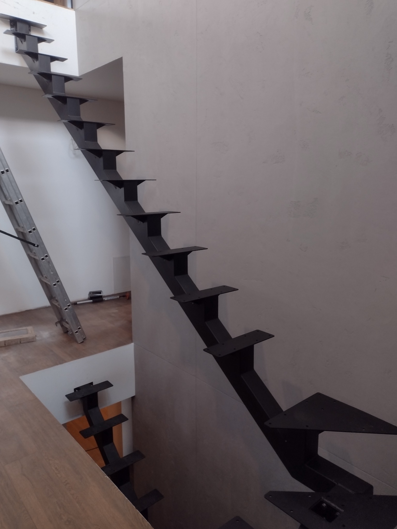 Surowe, czarne, metalowe schody bez poręczy, przymocowane do ściany w nowoczesnym wnętrzu. W tle drabina i narzędzia, co sugeruje etap montażu.