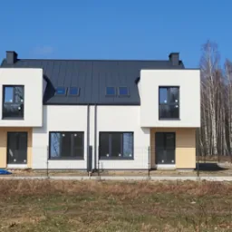 Nowoczesne dwukondygnacyjne bliźniaki z szarą dachówką i białymi elewacjami, widoczne okna i drzwi w ciemnych ramach, teren wokół ogrodzony siatką, w tle drzewa bez liści, błękitne niebo.