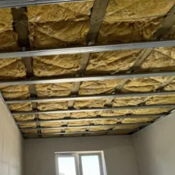 Montaż izolacji z wełny mineralnej w metalowym stelażu sufitu podwieszanego, widoczne fragmenty ścian i okno.