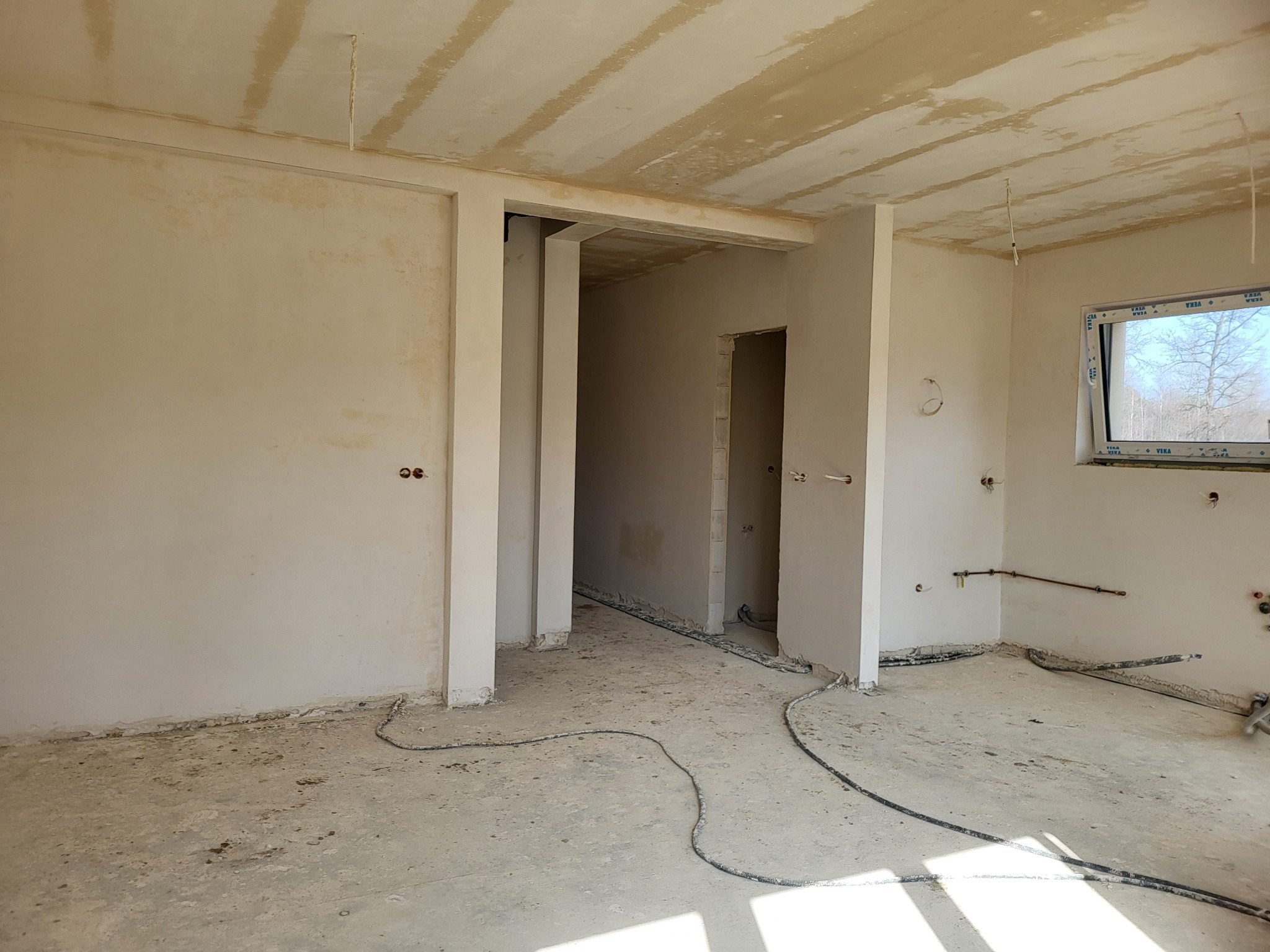 Wnętrze w stanie deweloperskim z surowymi ścianami, widocznymi instalacjami elektrycznymi i wodnymi, przygotowane do dalszych prac wykończeniowych, z oknem z widokiem na drzewa.