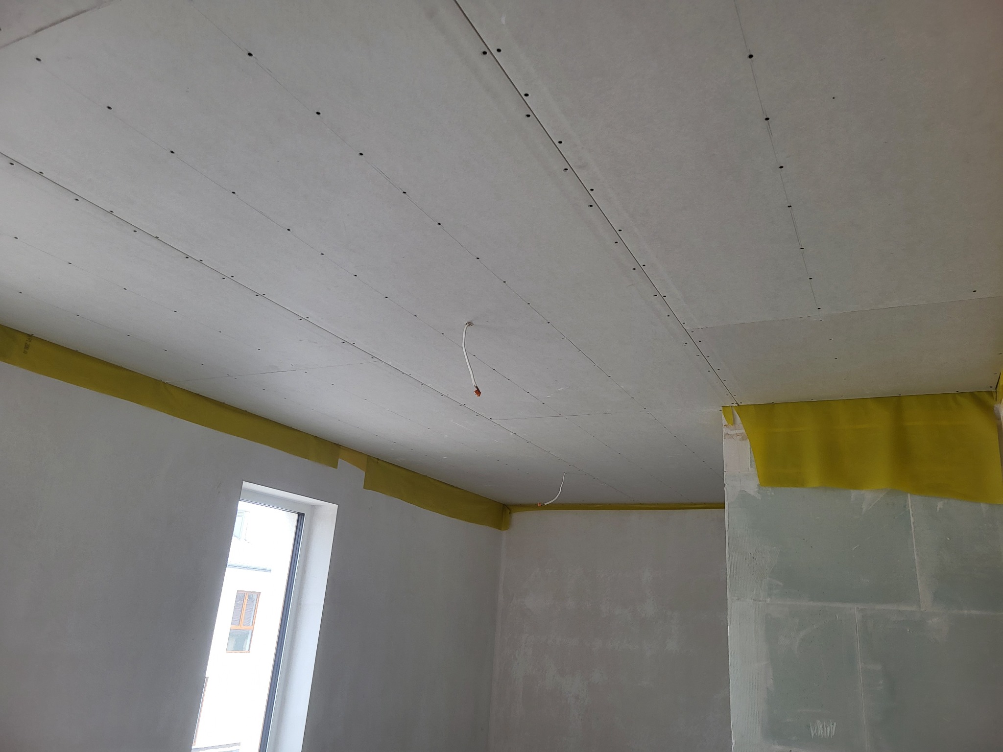 Montaż płyt kartonowo-gipsowych na suficie w pokoju, widoczne żółte taśmy zabezpieczające, surowy stan wykończenia ścian i wystający kabel.