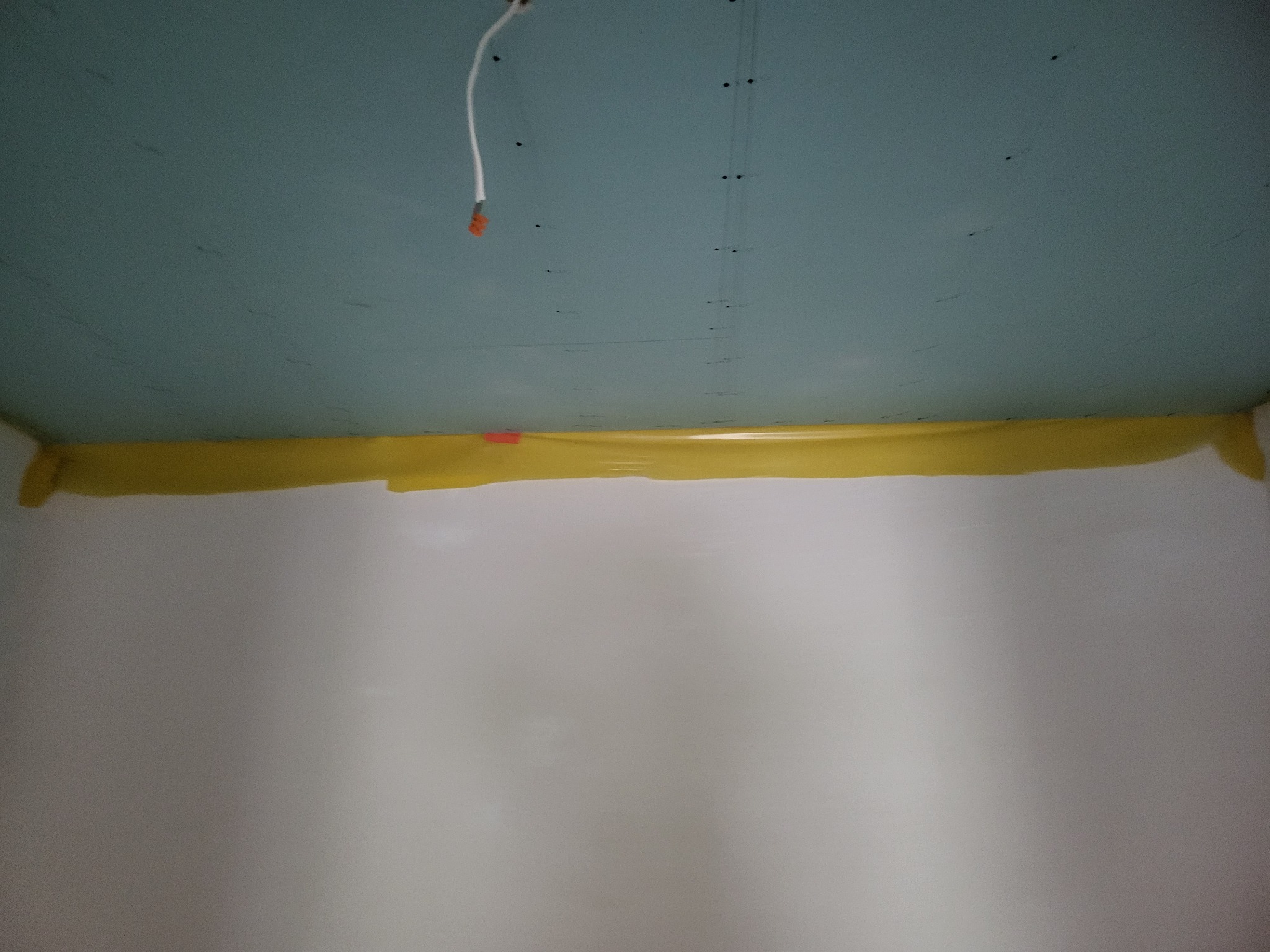 Świeżo zamontowany sufit z płyt kartonowo-gipsowych, zabezpieczony żółtą folią malarską przy ścianach, widoczne ślady po wkrętach i wystający kabel elektryczny.