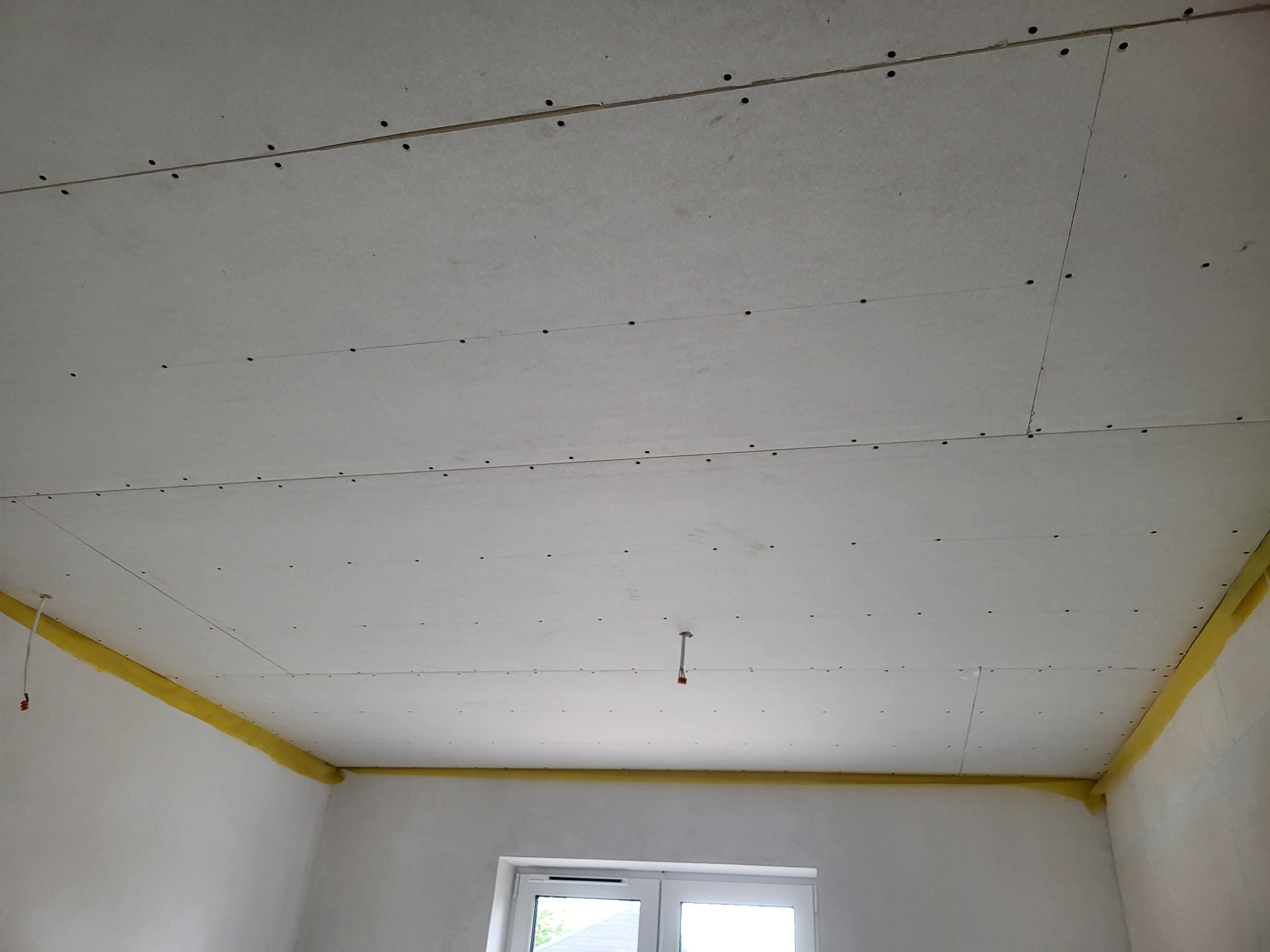 Sufit z płyt kartonowo-gipsowych, widoczne łączenia i wkręty, żółta taśma ochronna na styku ściany i sufitu, przygotowanie do dalszych prac wykończeniowych w pomieszczeniu z oknem.